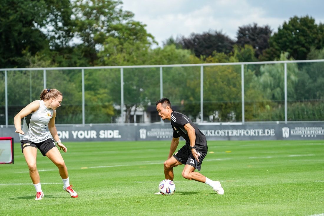 Zwei Personen trainieren auf einem Fußballfeld, während sie den Ball dribbeln. Im Hintergrund sind Bäume und eine Sporteinrichtung sichtbar.