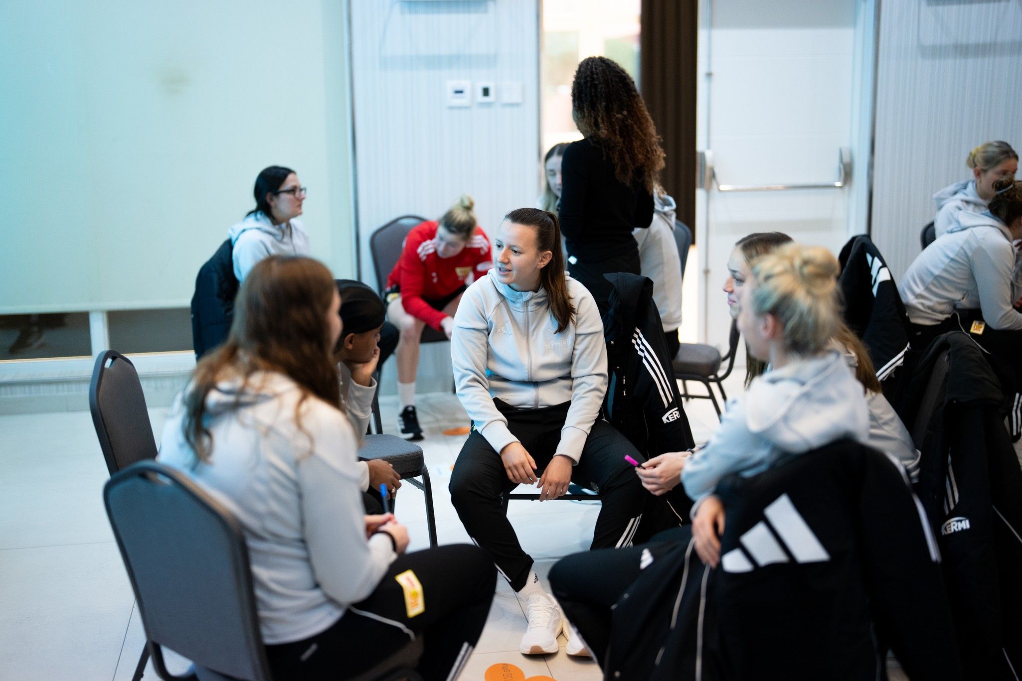 Eine Gruppe von Sportlerinnen in grauen Trainingsanzügen sitzt in einem Raum und unterhält sich.