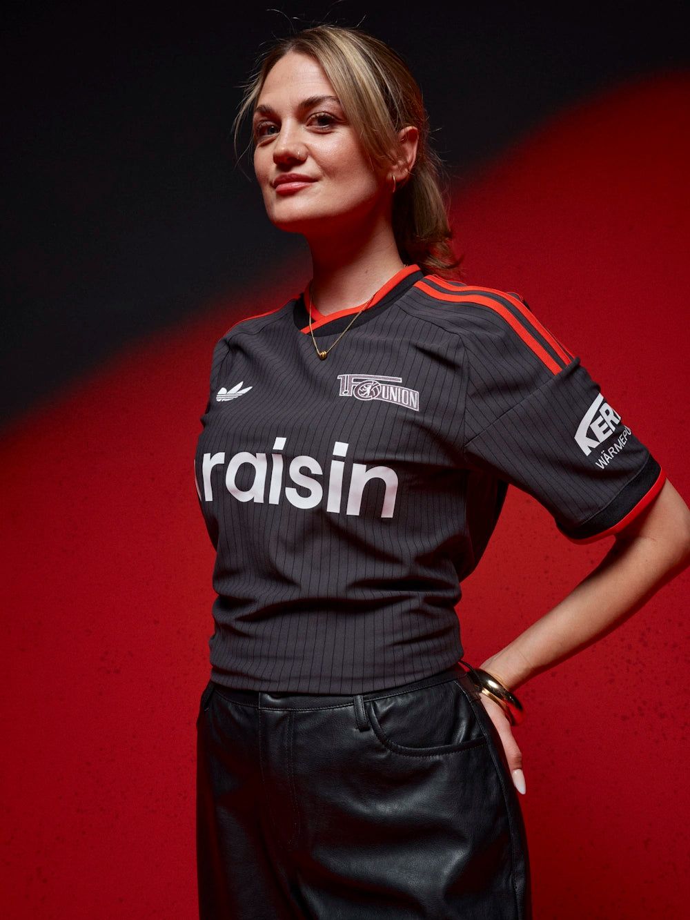 Frau in einem grau-schwarzen Fußballtrikot mit rotem Akzent und der Aufschrift "raisin", vor einem rot-schwarzen Hintergrund.