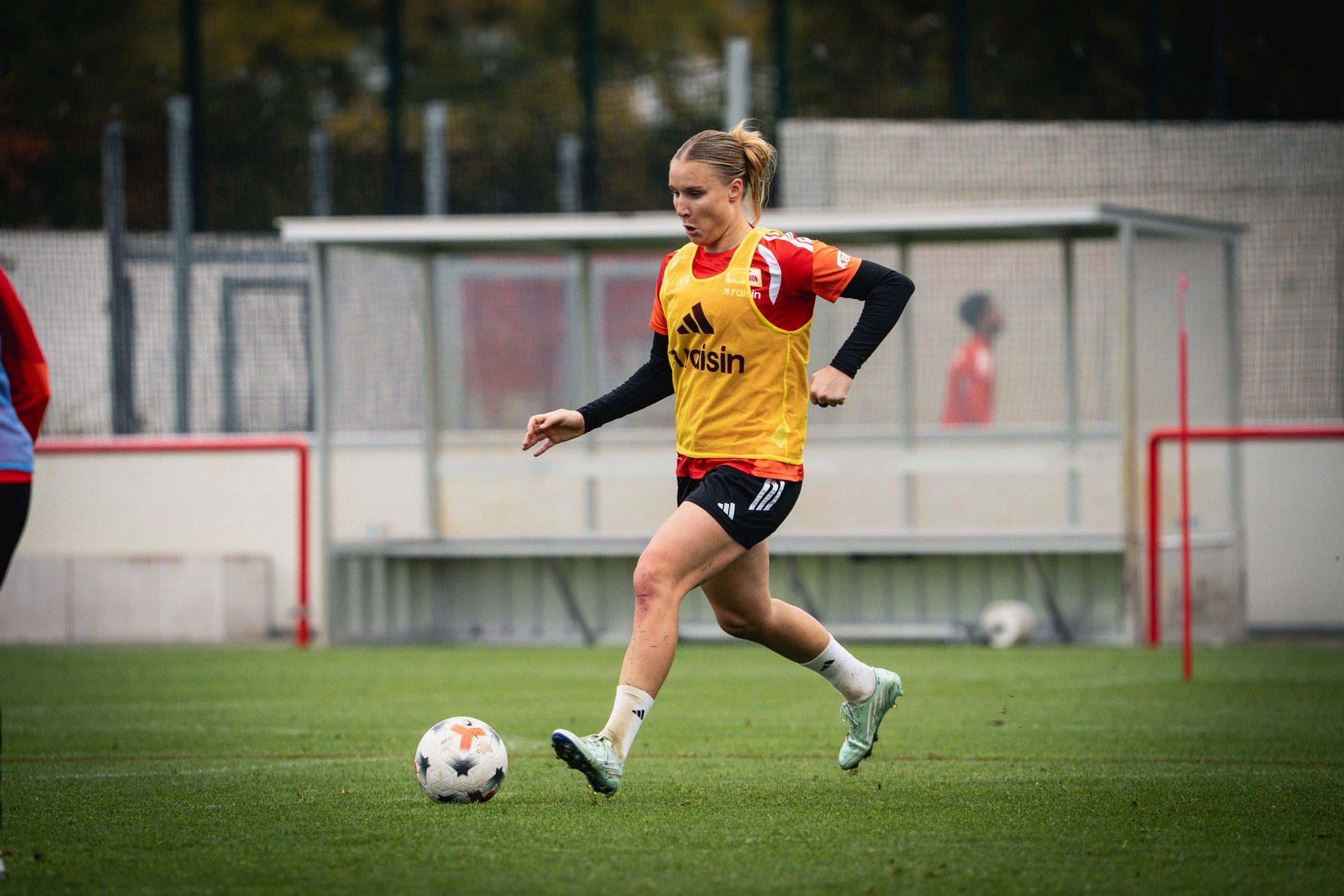 Eine Fußballspielerin in gelber Trainingsweste dribbelt den Ball auf dem Trainingsplatz.
