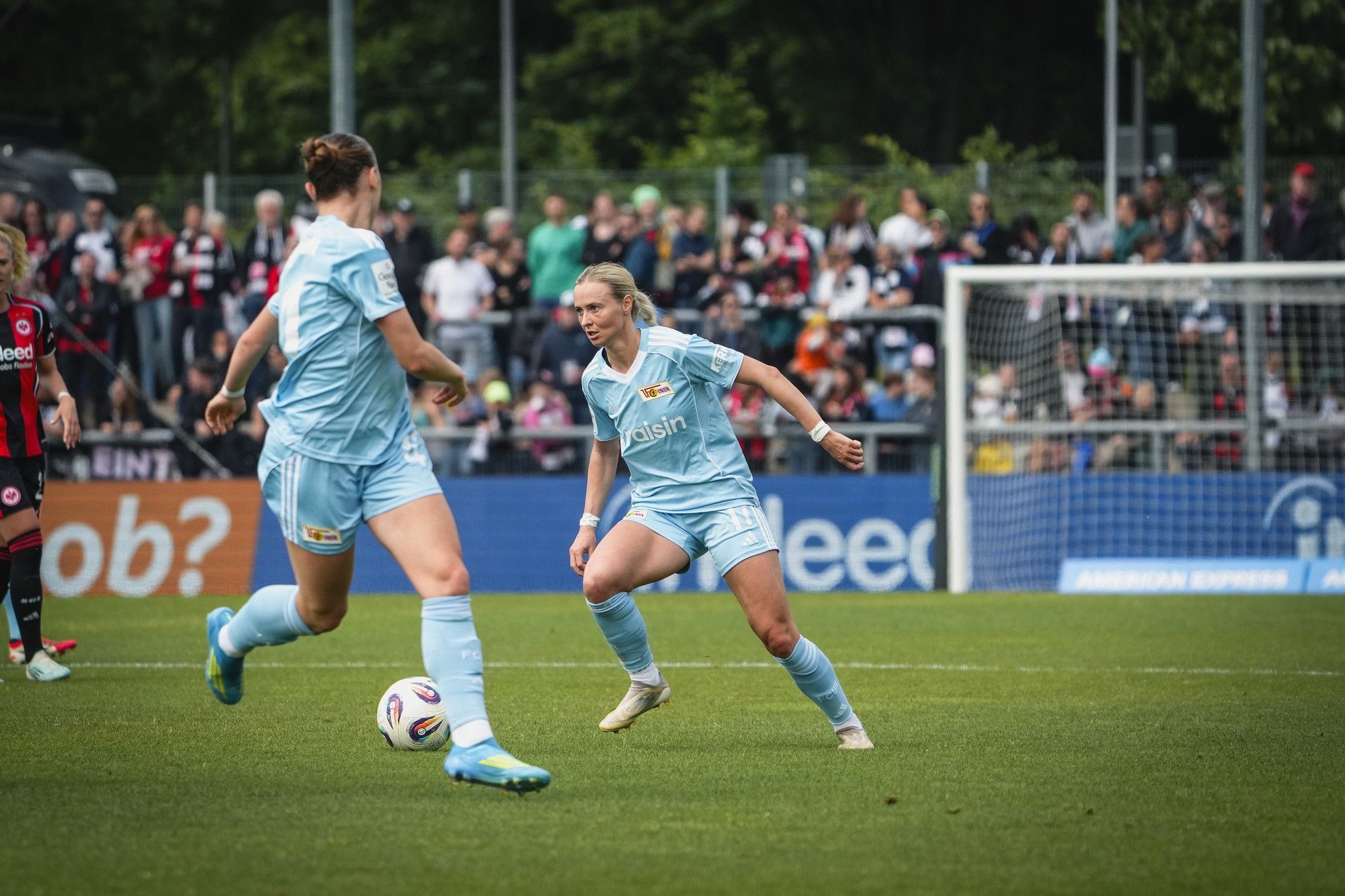 Zwei Fußballspielerinnen in hellblauen Trikots stehen auf dem Spielfeld, während Zuschauer im Hintergrund zuschauen.