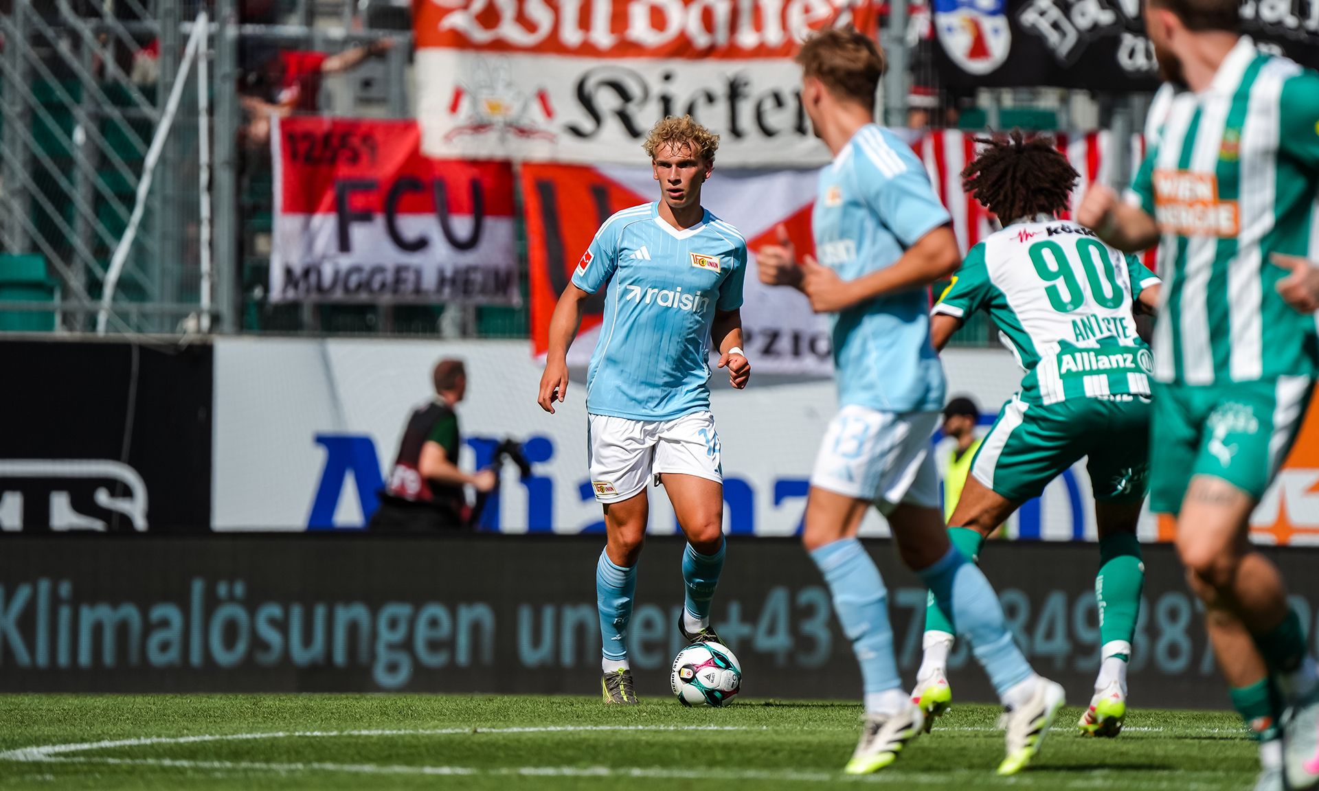 Spielszene im Fußball mit Spielern in blauen und grünen Trikots, umgeben von Fans und Flaggen im Hintergrund.