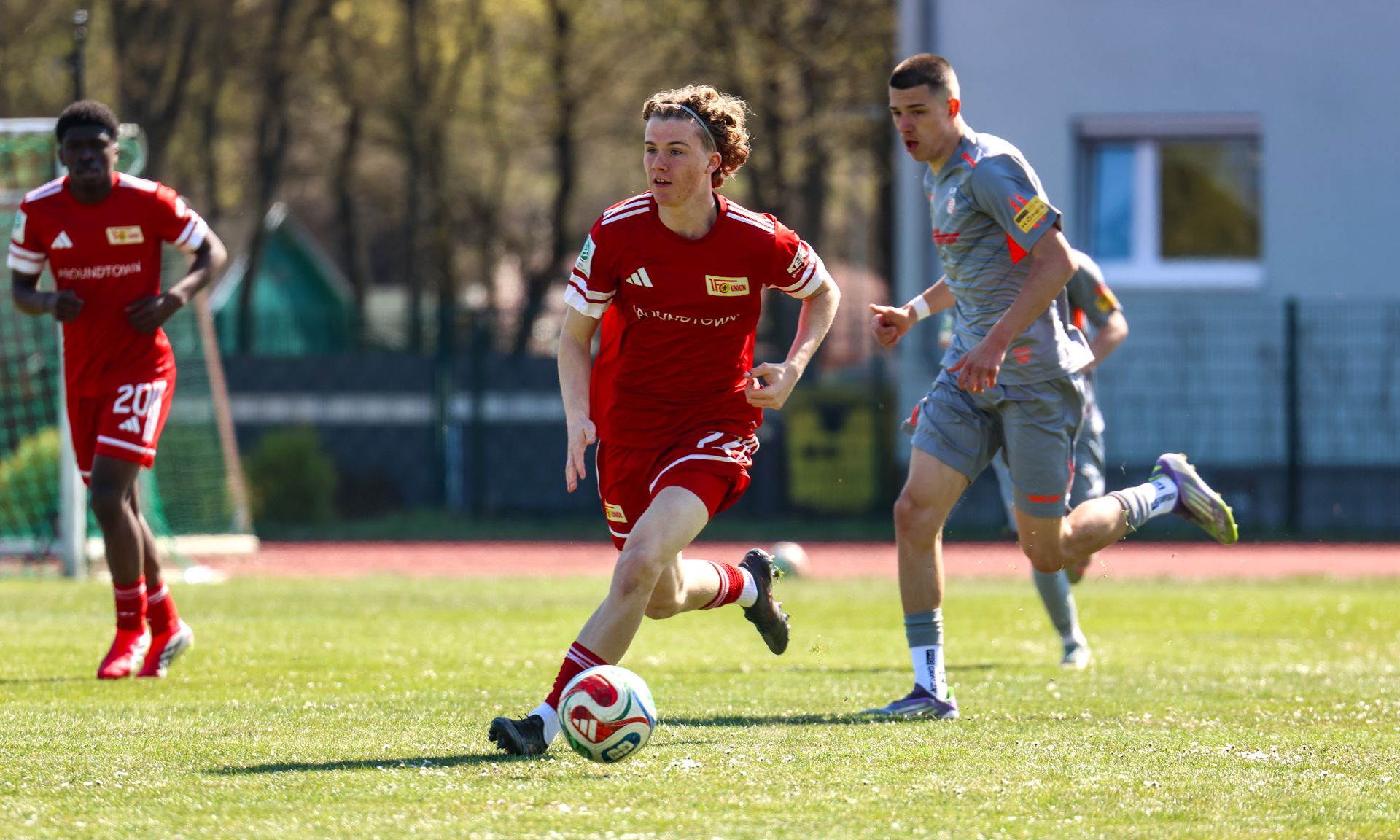 Ein Fußballspieler in rotem Trikot dribbelt den Ball, während ein Gegenspieler in grauem Trikot verfolgt. Hintergrund mit Bäumen.