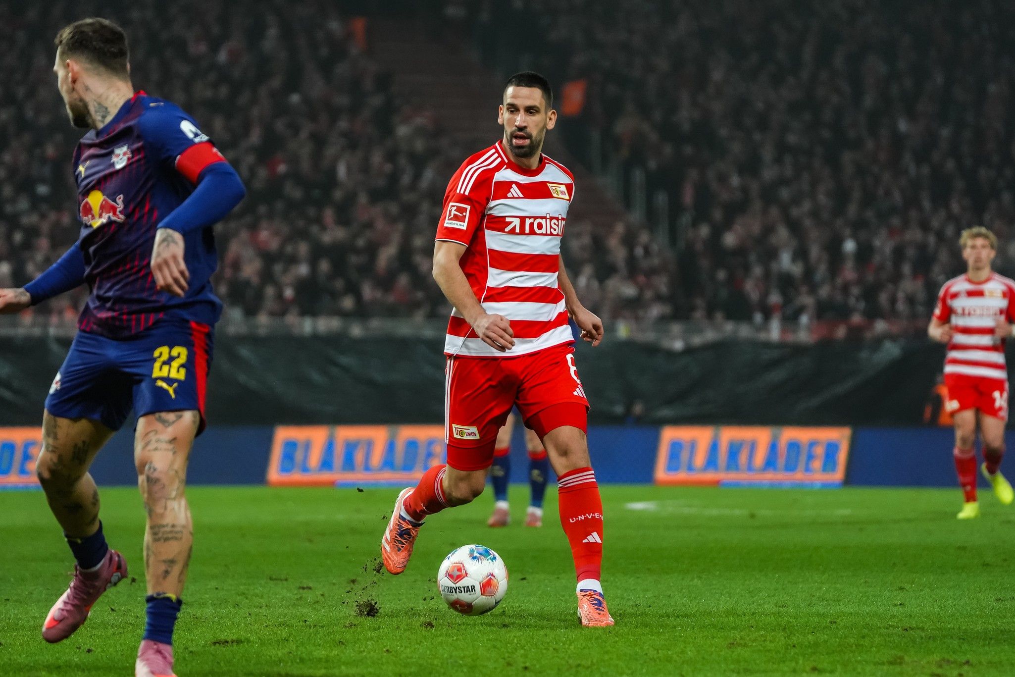 Ein Fußballspieler in rotem Trikot dribbelt den Ball, während ein Gegenspieler in blauem Trikot darauf reagiert. Stadionatmosphäre im Hintergrund.