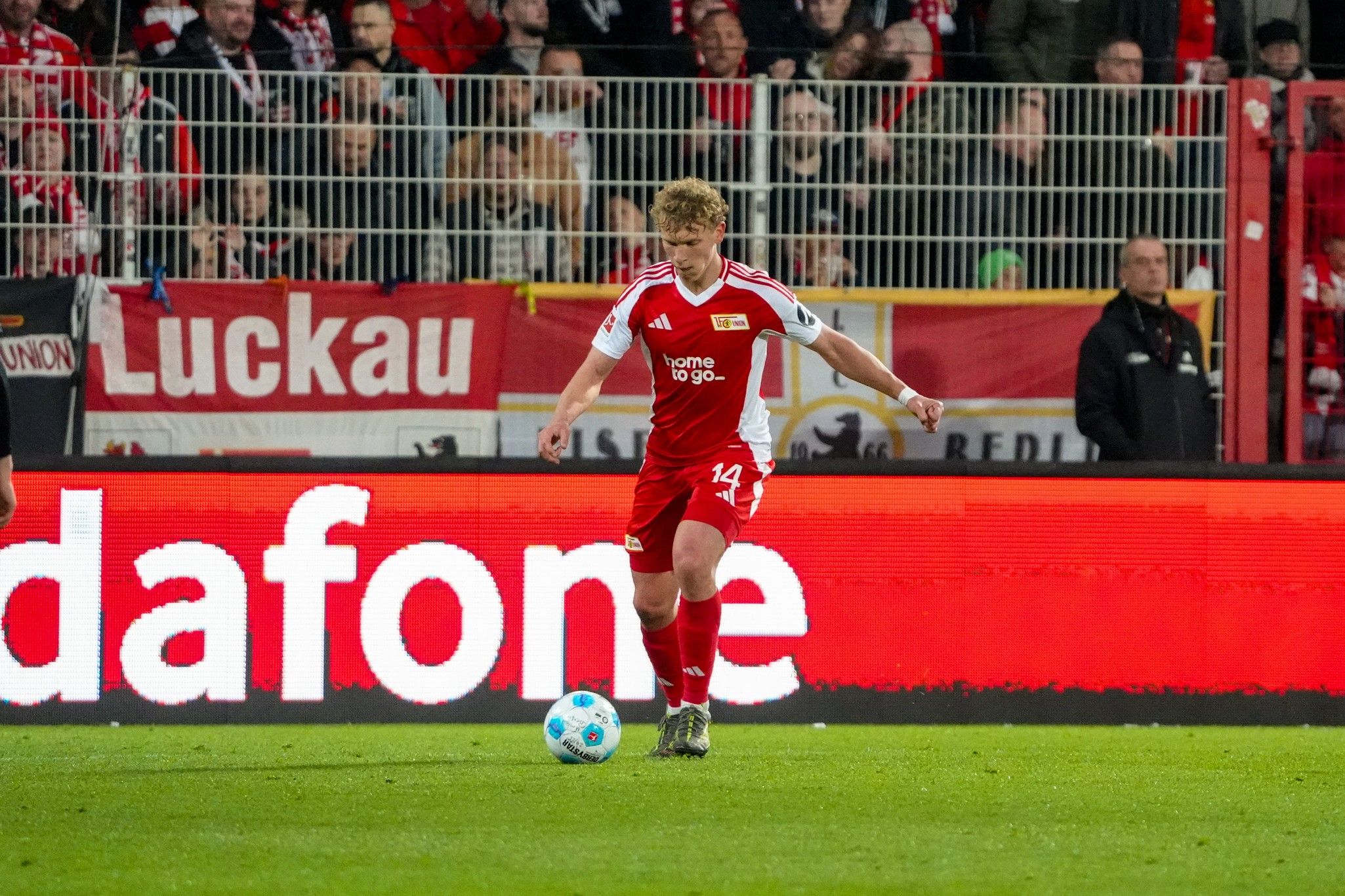 Spieler in rotem Trikot mit der Nummer 14 dribbelt den Ball auf dem Spielfeld, umgeben von jubelnden Fans.