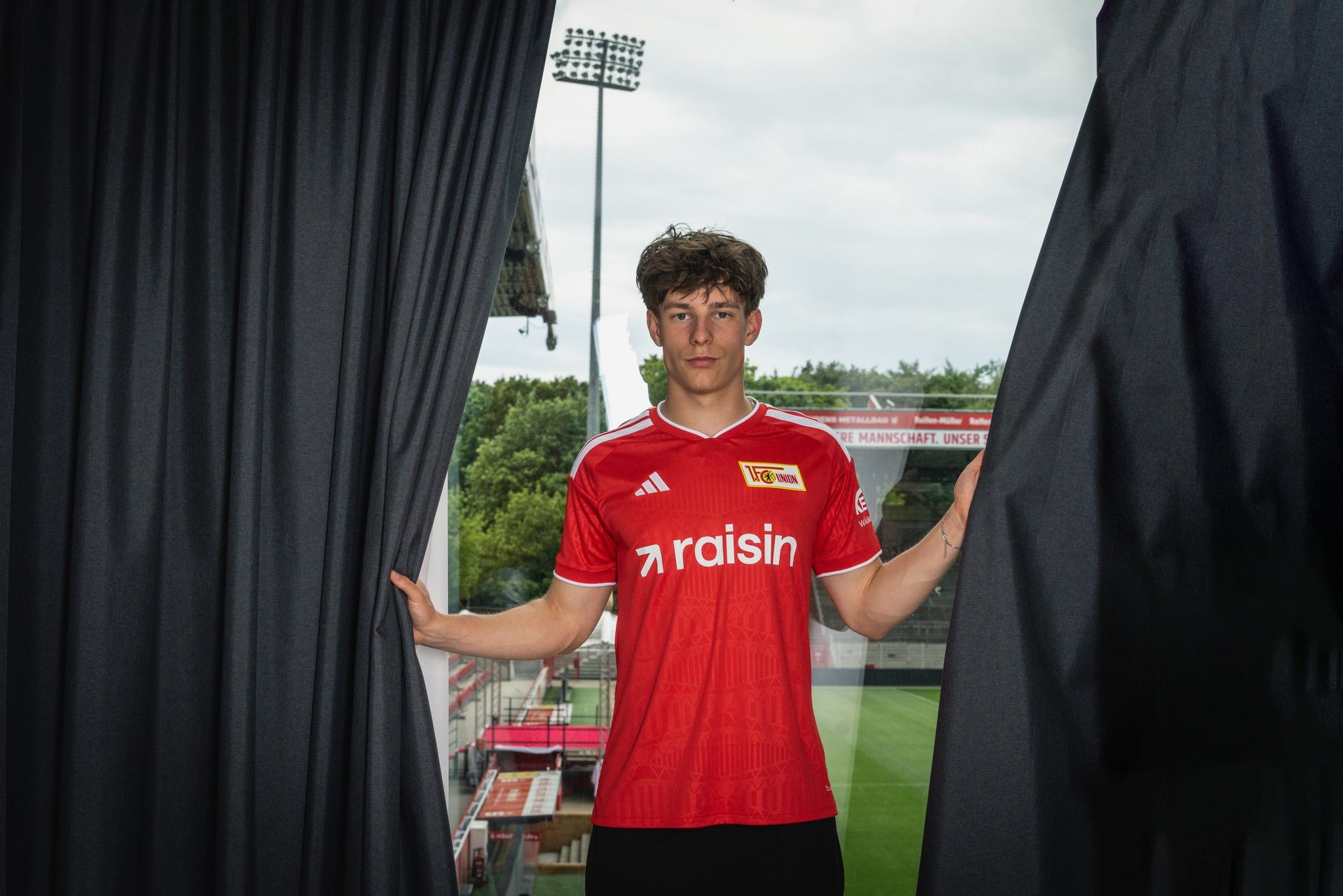 Ein junger Mann in einem roten Fußballtrikot steht zwischen Vorhängen und blickt auf ein Fußballfeld.