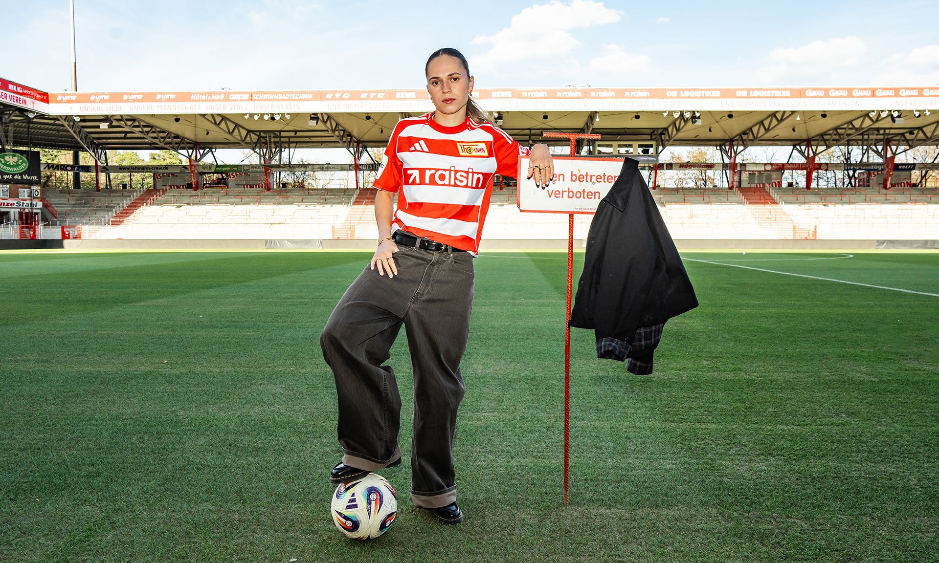Frau mit Fußball und gestreiftem Trikot steht auf dem Rasen eines Stadions neben einem Eckfahne.