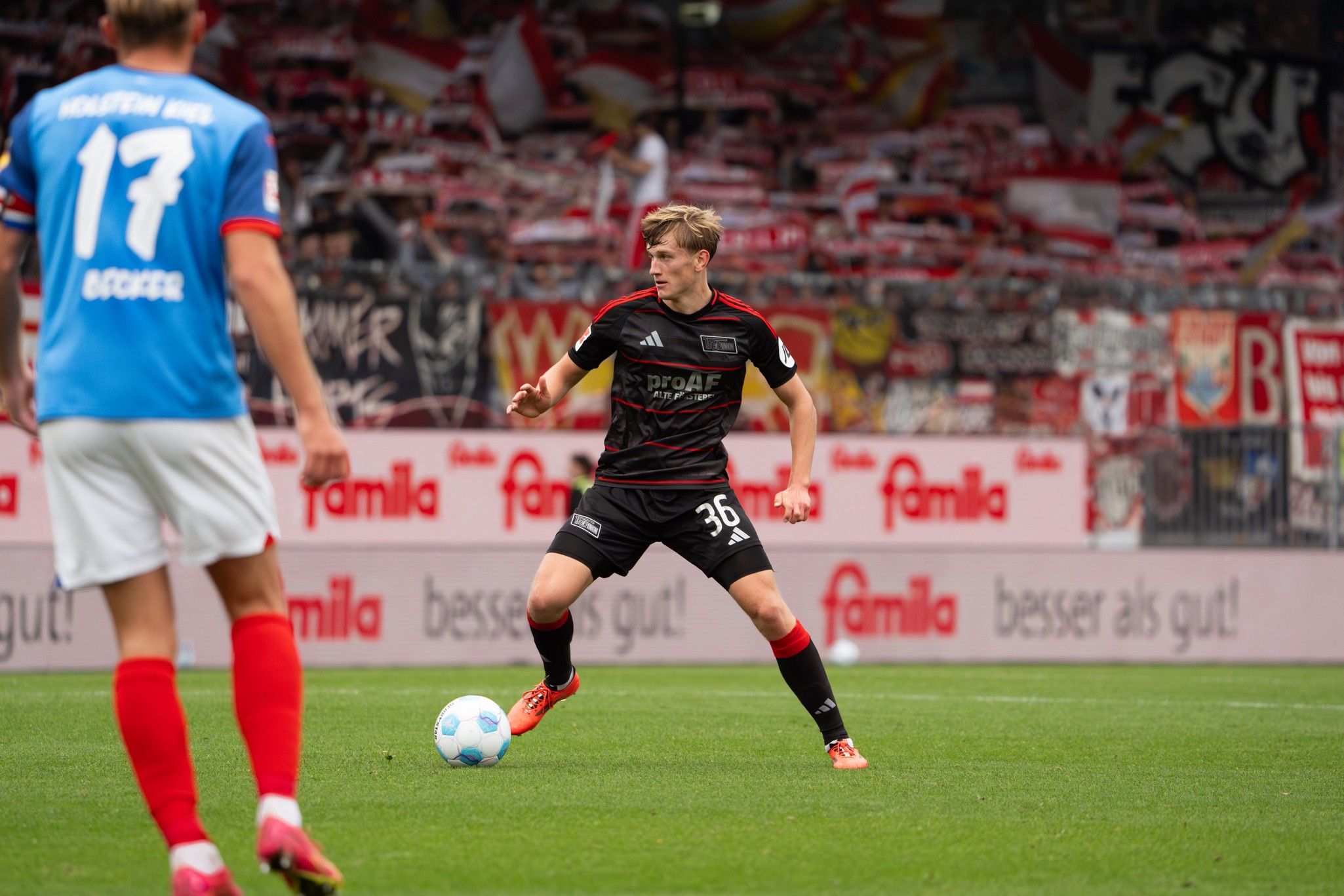 Ein Fußballspieler mit der Nummer 36 dribbelt den Ball, während ein anderer Spieler in roter und blauer Kleidung ihm gegenübersteht.