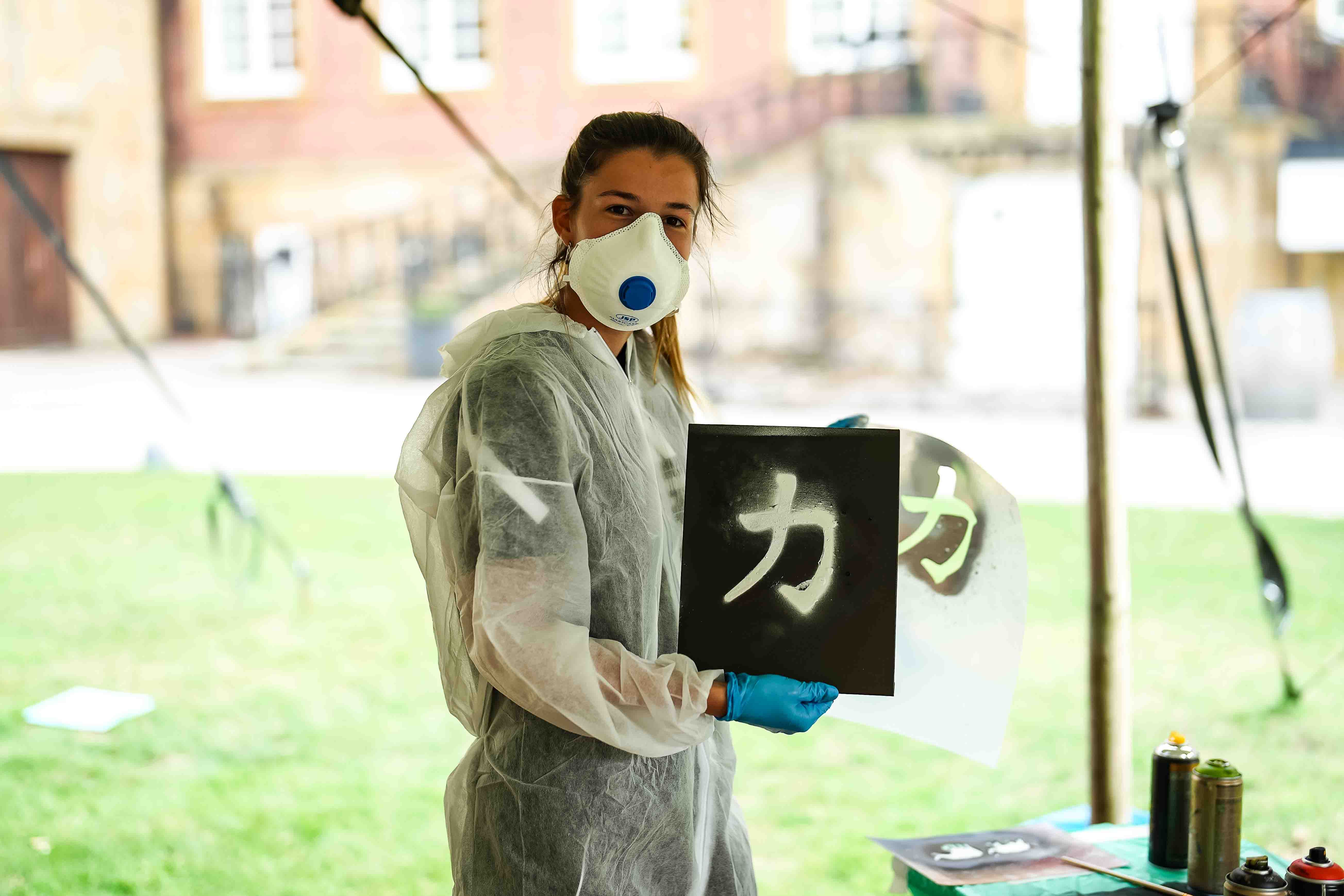 Eine Person in Schutzkleidung und Maske hält ein schwarzes Plakat mit einer weißen Schriftzeichen-Illumination in der Hand.
