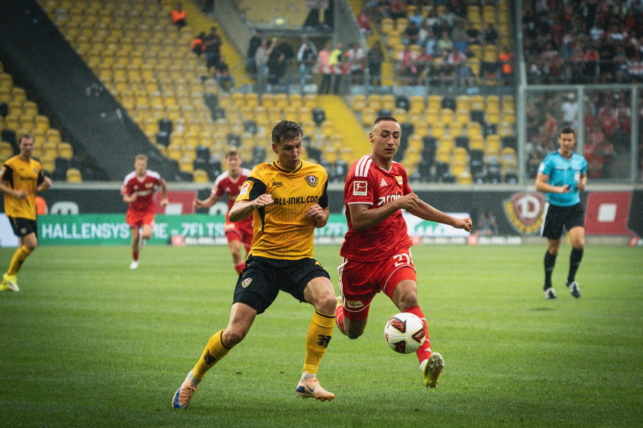 Ein Fußballspiel bei Regen, in dem ein gelb gekleideter Spieler einen roten Spieler mit Ball verfolgt. Zuschauer im Hintergrund.
