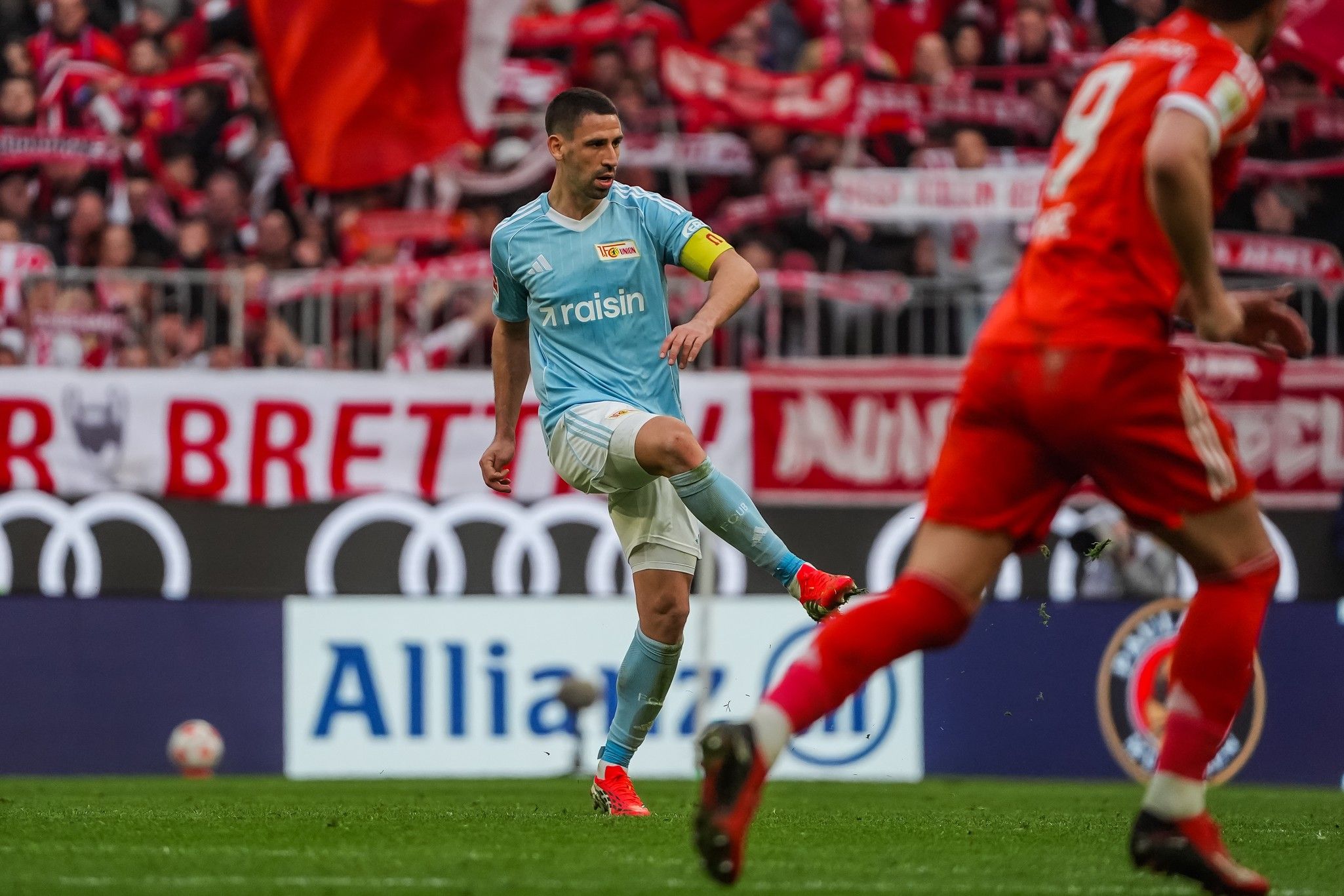 Ein Fußballspieler in hellblauer Torwartuniform schießt den Ball, während ein anderer Spieler in roter Uniform auf ihn zuläuft.