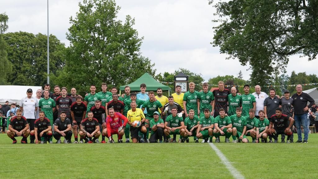 Gruppenfoto von Fußballspielern und Trainern auf einem Rasenfeld, umgeben von Zuschauern und Bäumen.