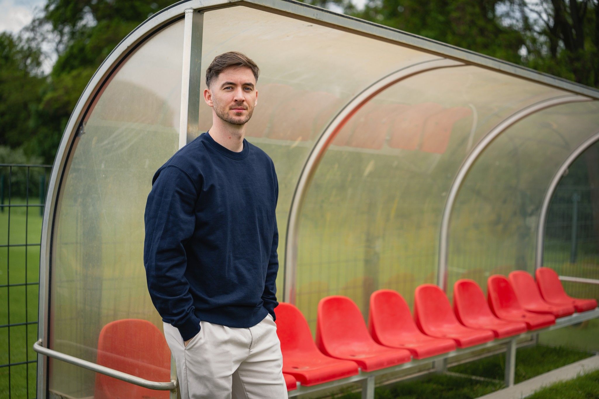 Mann in Blau steht vor einer Trainerbank mit roten Sitzen auf einem Sportplatz. Hintergrund mit grüner Landschaft.