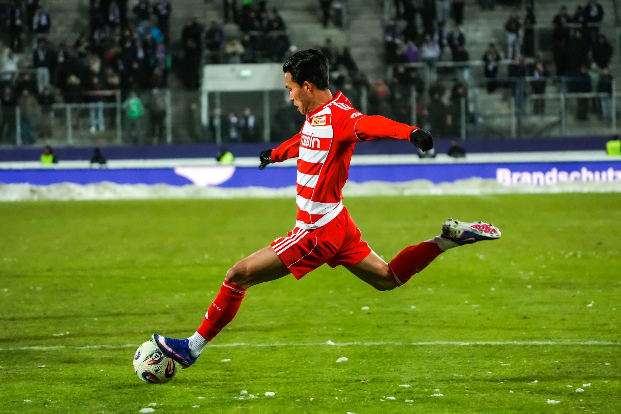 Ein Fußballspieler in rotem Trikot schießt den Ball auf einem schneebedeckten Spielfeld. Fans sind im Hintergrund sichtbar.