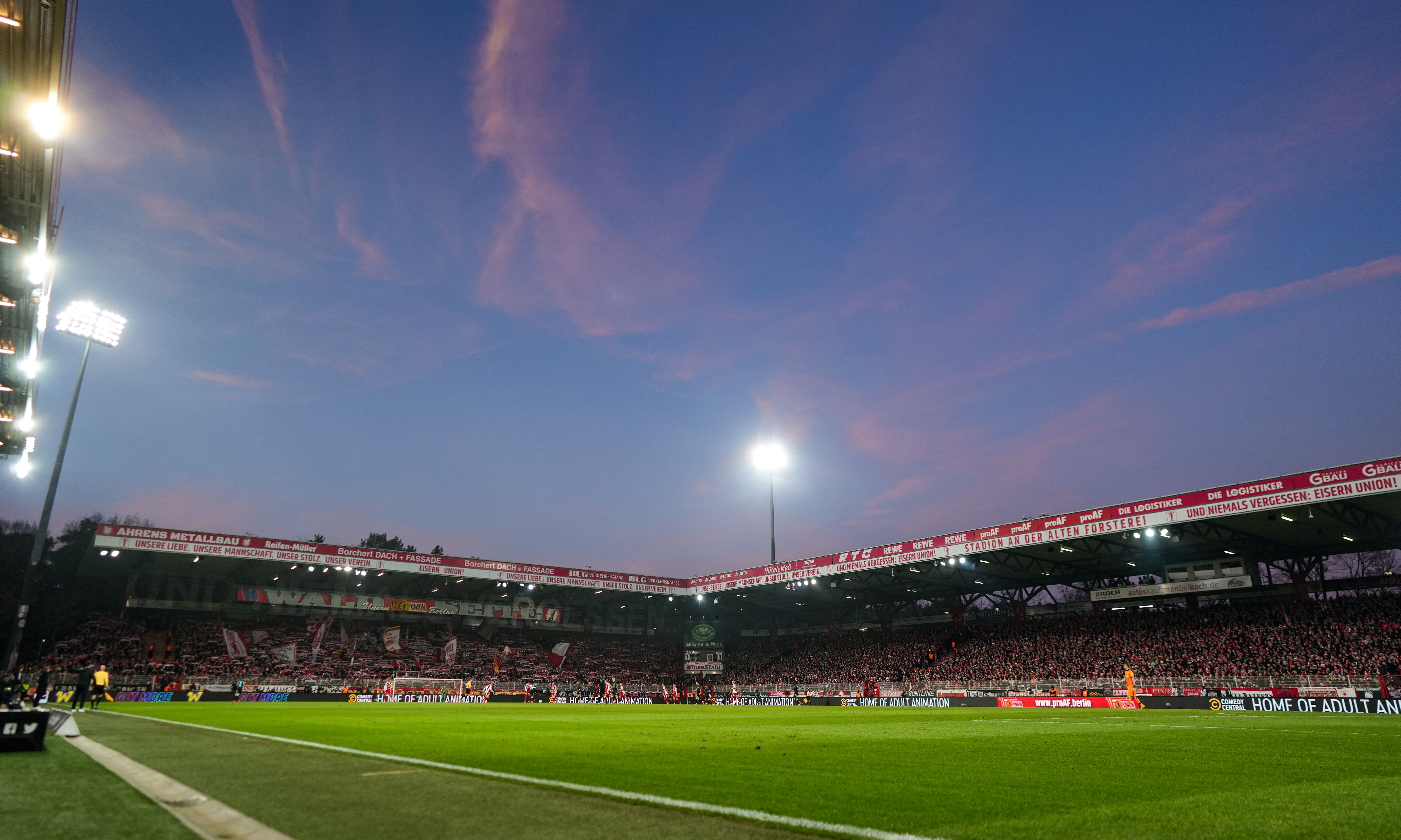 Stadionansicht bei Dämmerung mit rötlichem Himmel, beleuchteten Tribünen und Zuschauern auf der Tribüne.