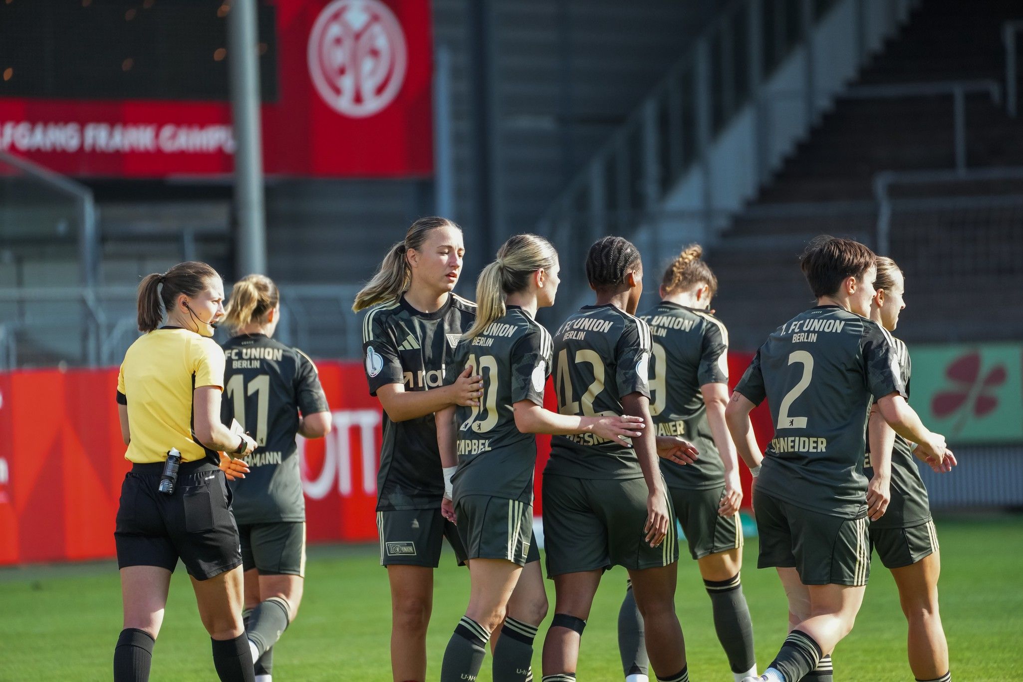 Frauenfußballmannschaft in grünen Trikots, die auf dem Spielfeld zusammenkommt, während eine Schiedsrichterin beobachtet.