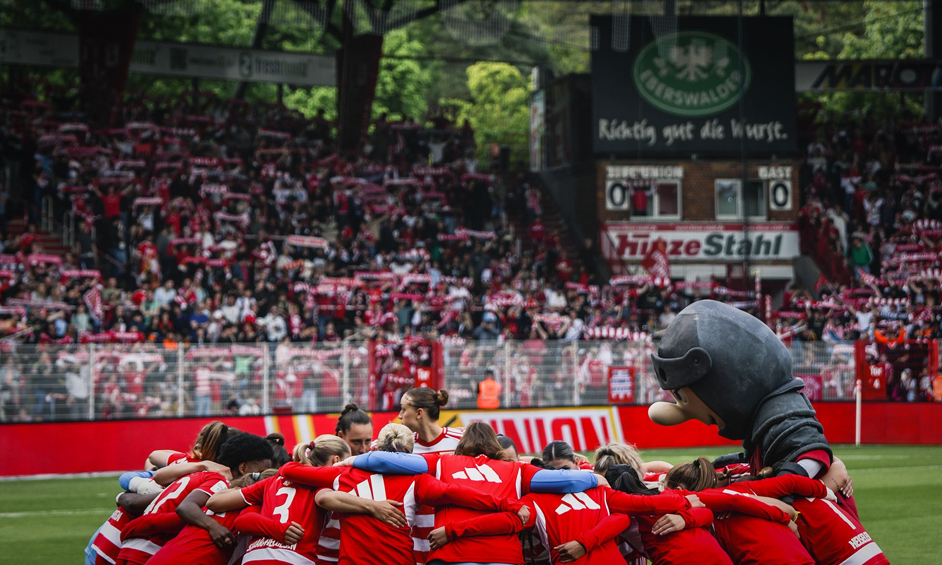 Mitspieler in roten Trikots umarmen sich im Kreis, während zahlreiche Fans mit Schals im Hintergrund jubeln. Ein Maskottchen ist ebenfalls zu sehen.