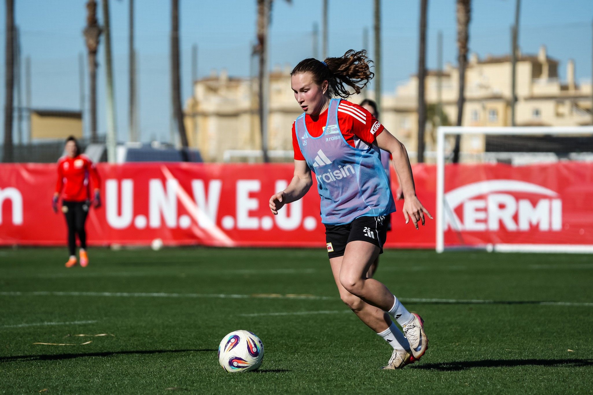 Eine Fußballspielerin in Trikots läuft auf einem grünen Feld während eines Trainings. Im Hintergrund sind Palmen und Banner zu sehen.