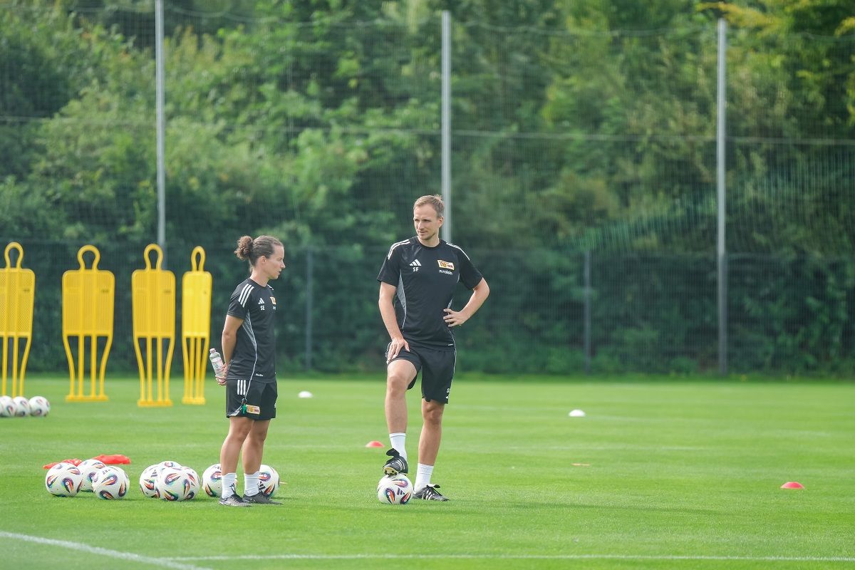 Zwei Trainer stehen auf einem Fußballplatz, umgeben von Fußbällen und Trainingshilfen, während sie sich unterhalten.