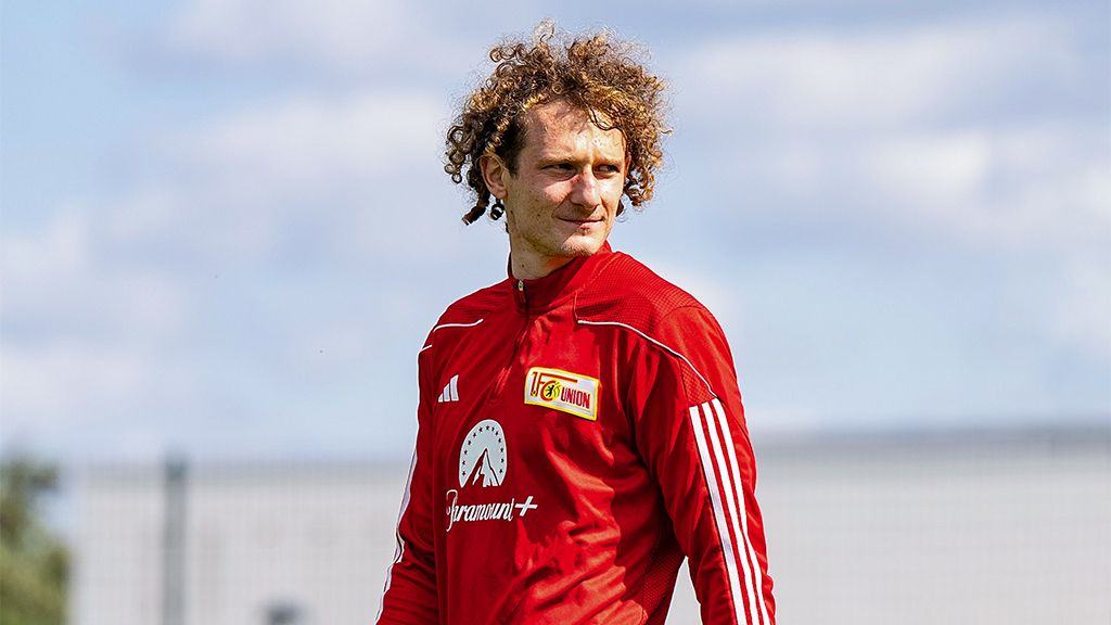 Ein Fußballspieler mit lockigen Haaren trägt ein rotes Trainingsoutfit und schaut nachdenklich in die Ferne.