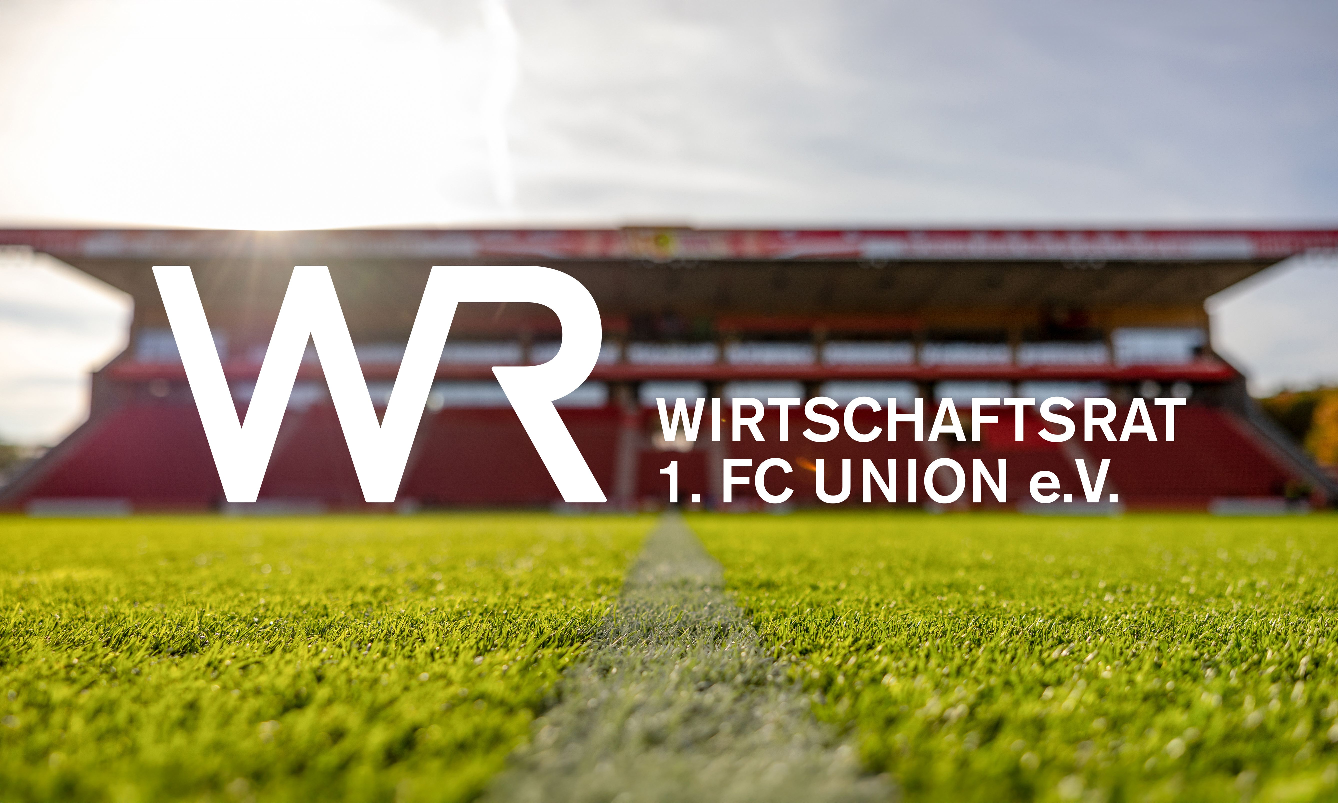 Logo des Wirtschaftsrats des 1. FC Union e.V. vor einem Fußballstadion mit grüner Rasenfläche.
