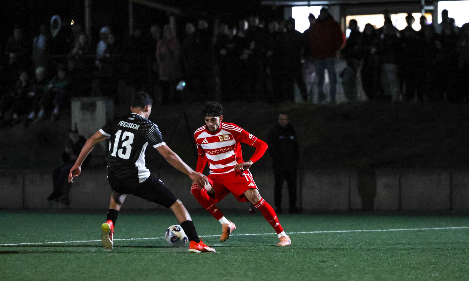 Zwei Fußballspieler im Wettkampf auf einem dunklen Spielfeld, umgeben von zuschauenden Fans.