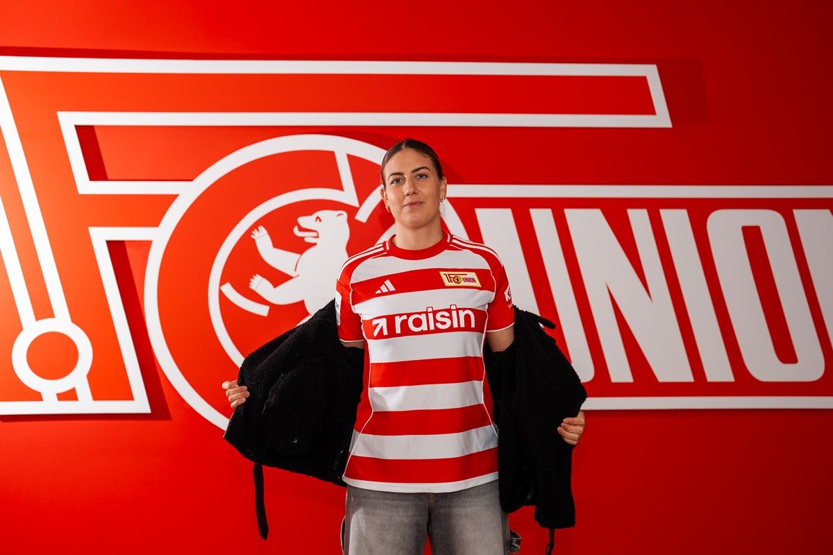 Eine Person in einem rot-weißen Fußballtrikot steht vor einem Logo des FC Union Berlin auf rotem Hintergrund.