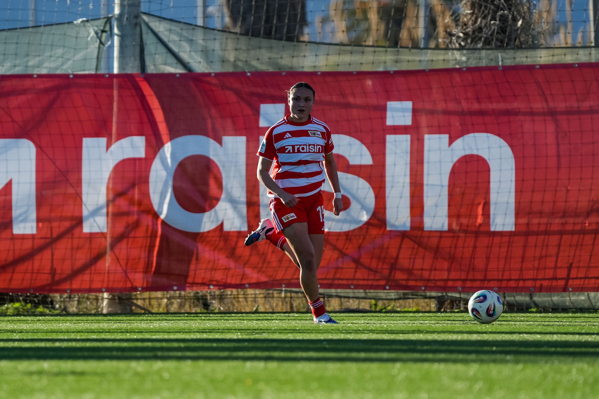 Spielerin in rotem Fußballtrikot mit weißen Streifen läuft auf dem Spielfeld, im Hintergrund eine große Werbebande.