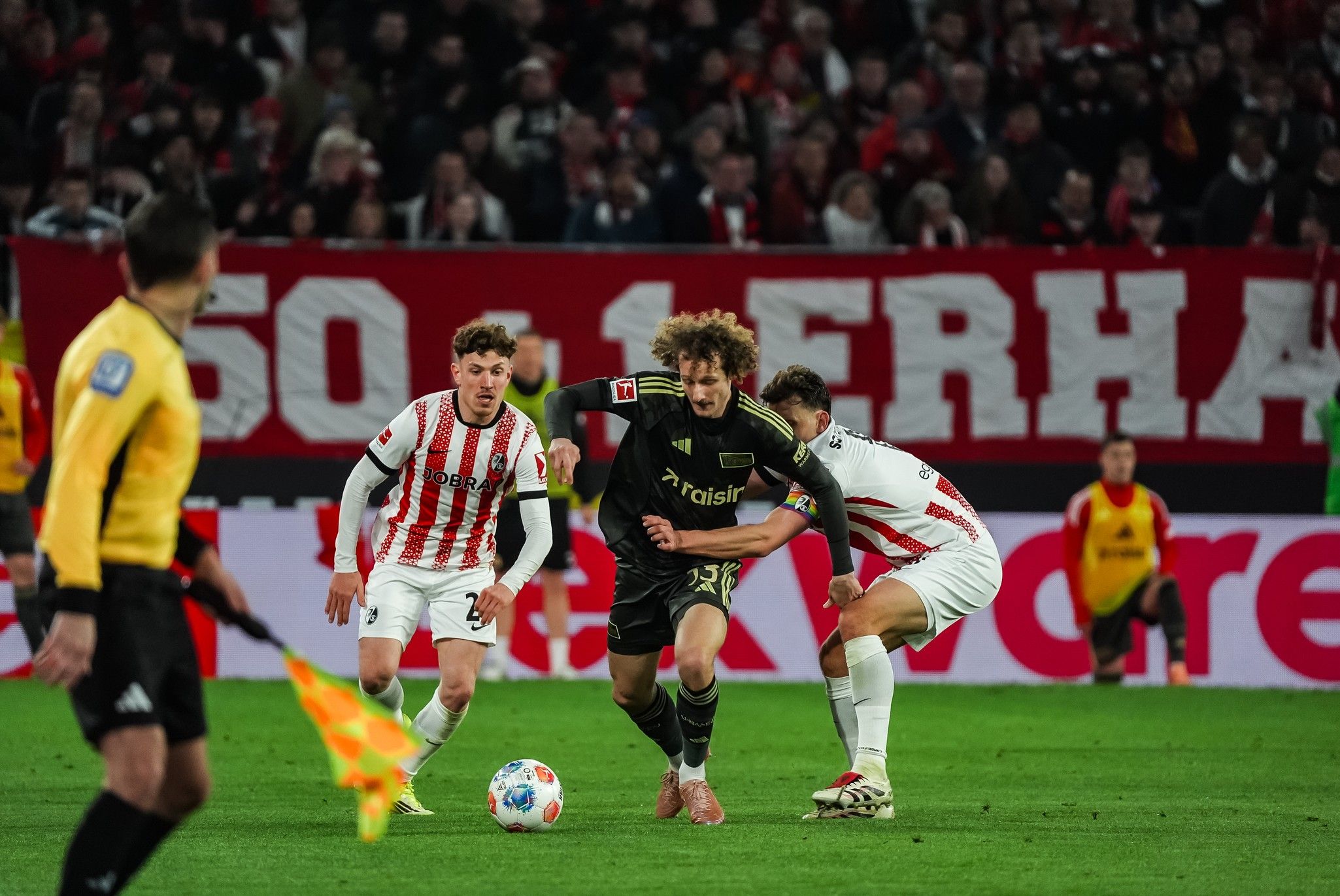 Ein Fußballspieler dribbelt den Ball, während zwei Verteidiger ihn verfolgen, im Hintergrund eine große Zuschauertribüne.