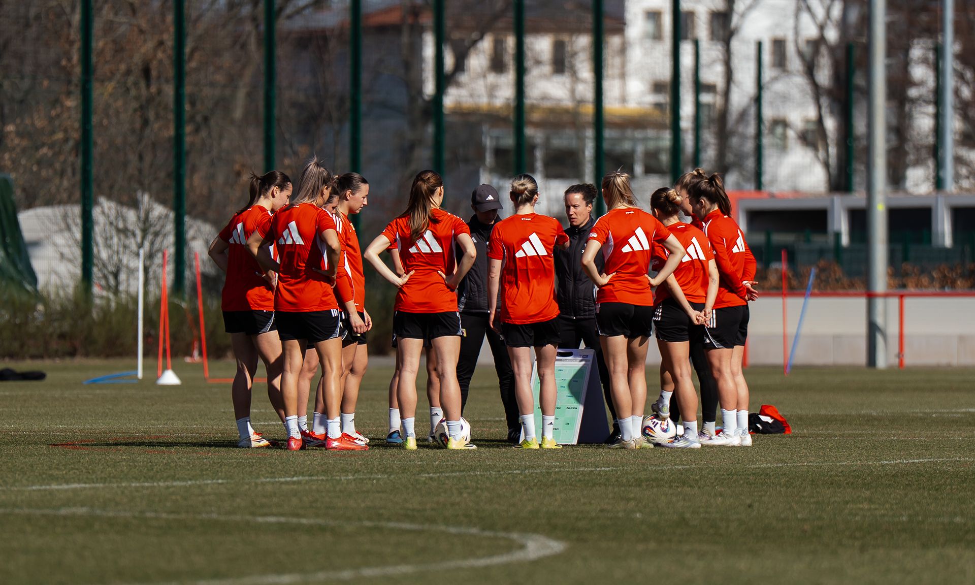 Eine Gruppe von Fußballspielerinnen in roten Trikots versammelt sich um einen Trainer auf einem grünen Trainingsfeld.