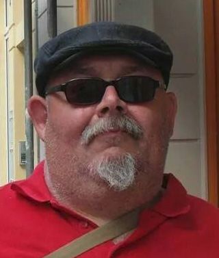 Mann mit Brille und Kappe, trägt ein rotes Shirt und steht vor einer hellen Wand.