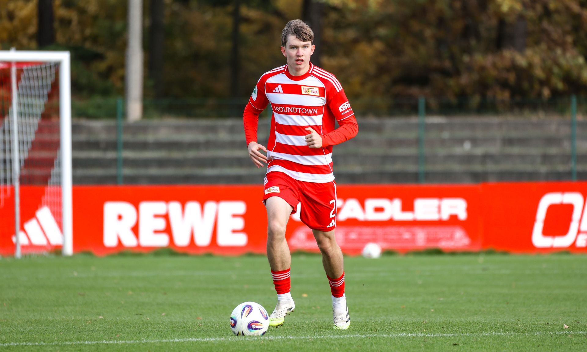 Spieler in rotem Trikot mit Streifen dribbelt den Ball auf einem Fußballfeld. Hintergrund zeigt Werbeflächen und Bäume.