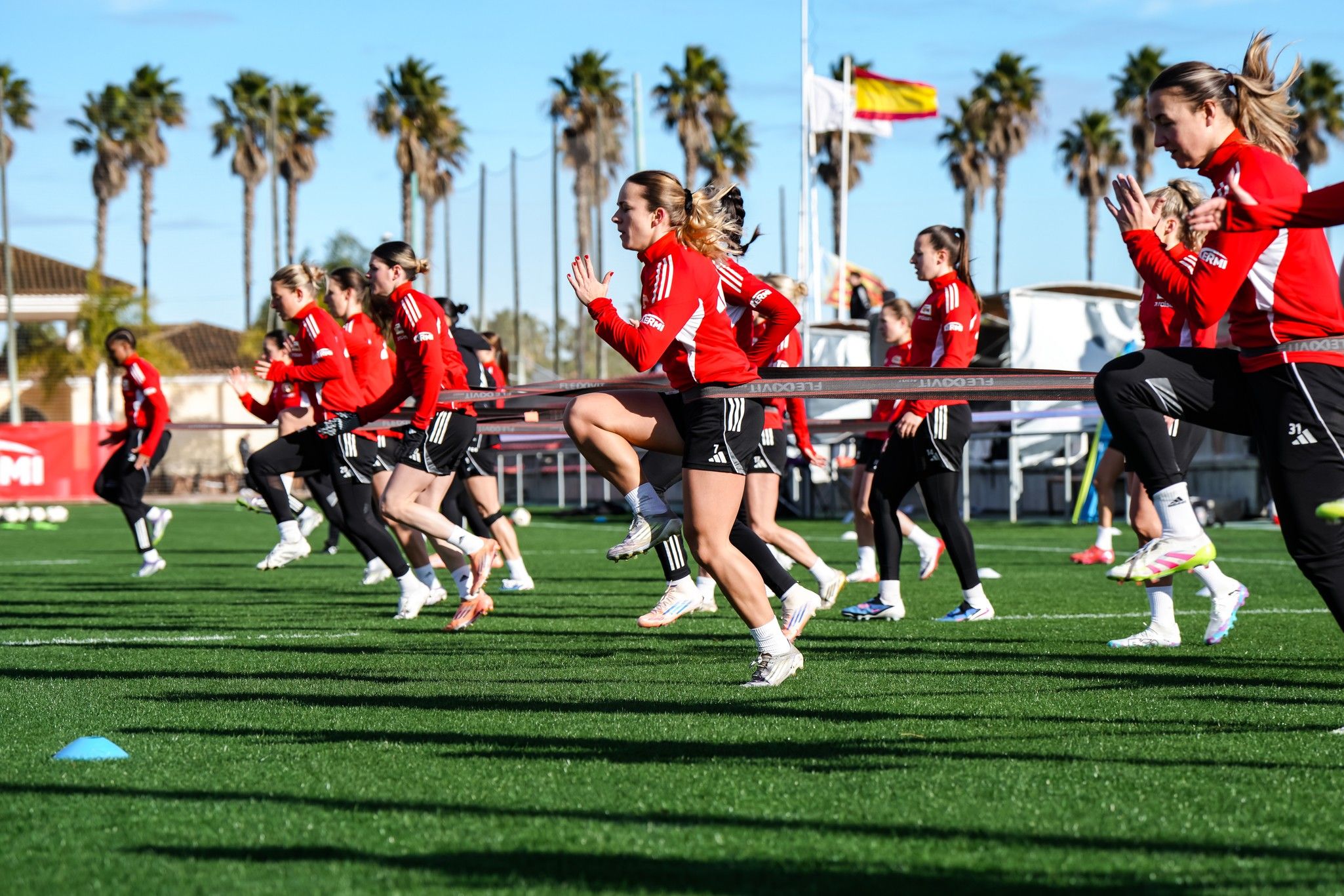 Mannschaft von Spielerinnen in roten Trainingsanzügen beim Sprinttraining auf einem grünen Kunstrasenplatz.