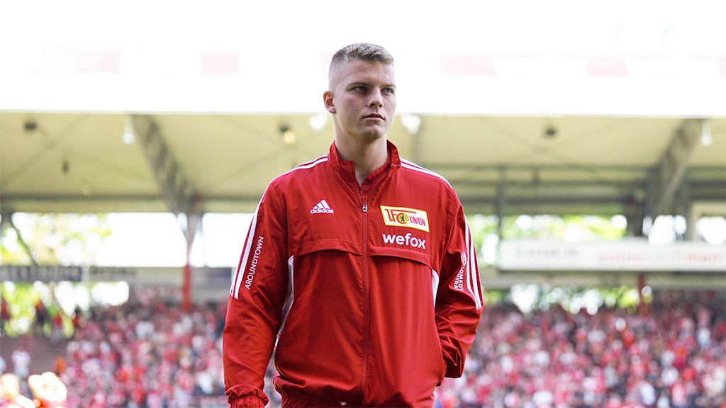 Ein junger Fußballspieler in roter Trainingsjacke steht auf einem Stadionfeld, im Hintergrund ist die Menge zu sehen.