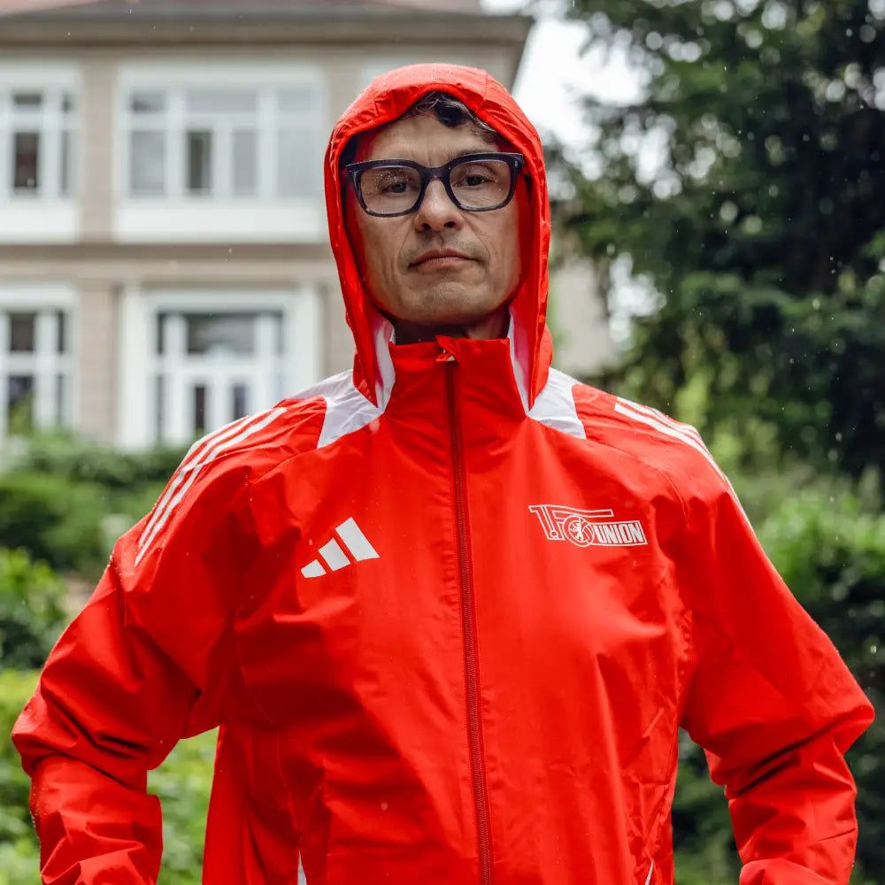 Eine Person trägt eine rote Sportjacke mit Kapuze. Sie steht draußen. Im Hintergrund sind grüne Wiesen und ein Gebäude.