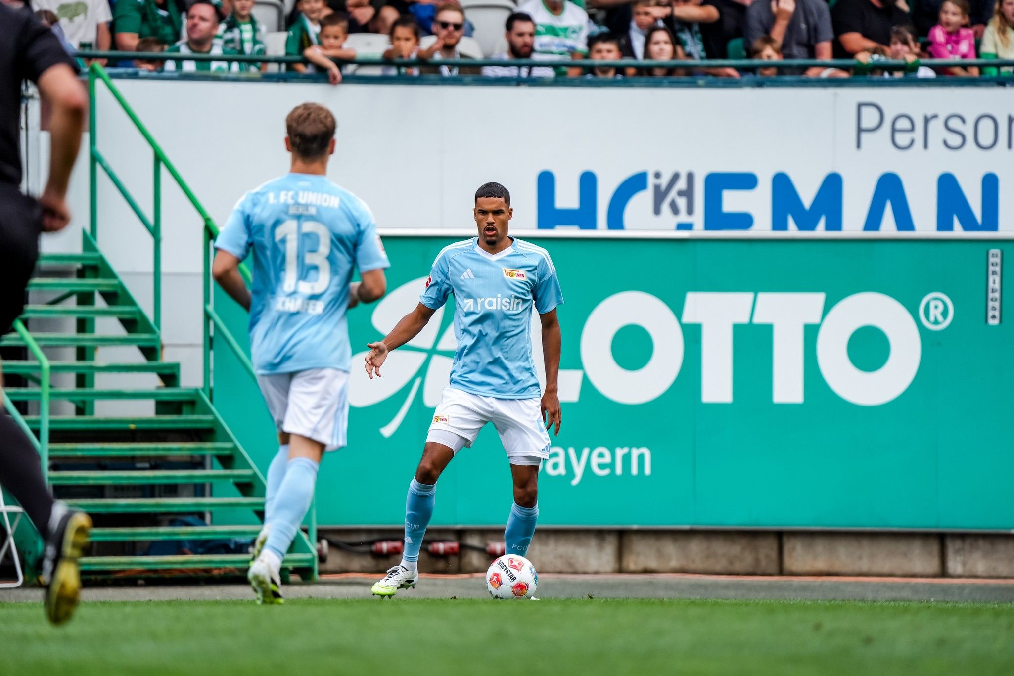 Ein Fußballspieler in hellblauem Trikot steht am Ball, im Hintergrund stehen Zuschauer auf einer Tribüne.
