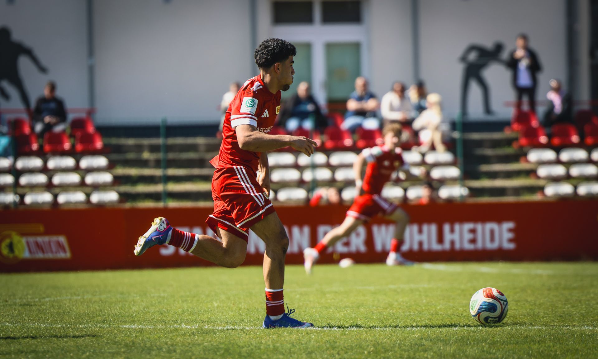 Ein Fußballspieler in rotem Trikot läuft auf dem Spielfeld, während ein weiterer Spieler im Hintergrund aktiv ist.