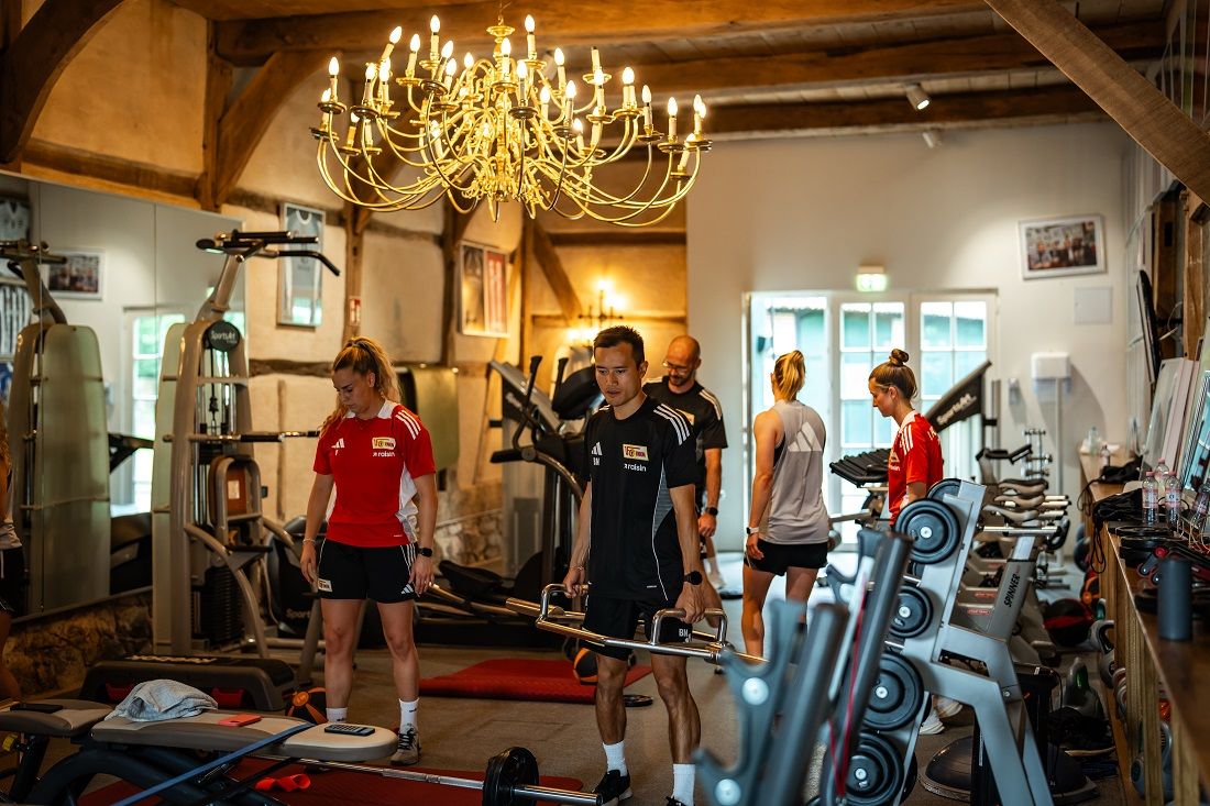 Personen trainieren in einem gut ausgestatteten Fitnessstudio mit verschiedenen Trainingsgeräten und einer eleganten Deckenleuchte.