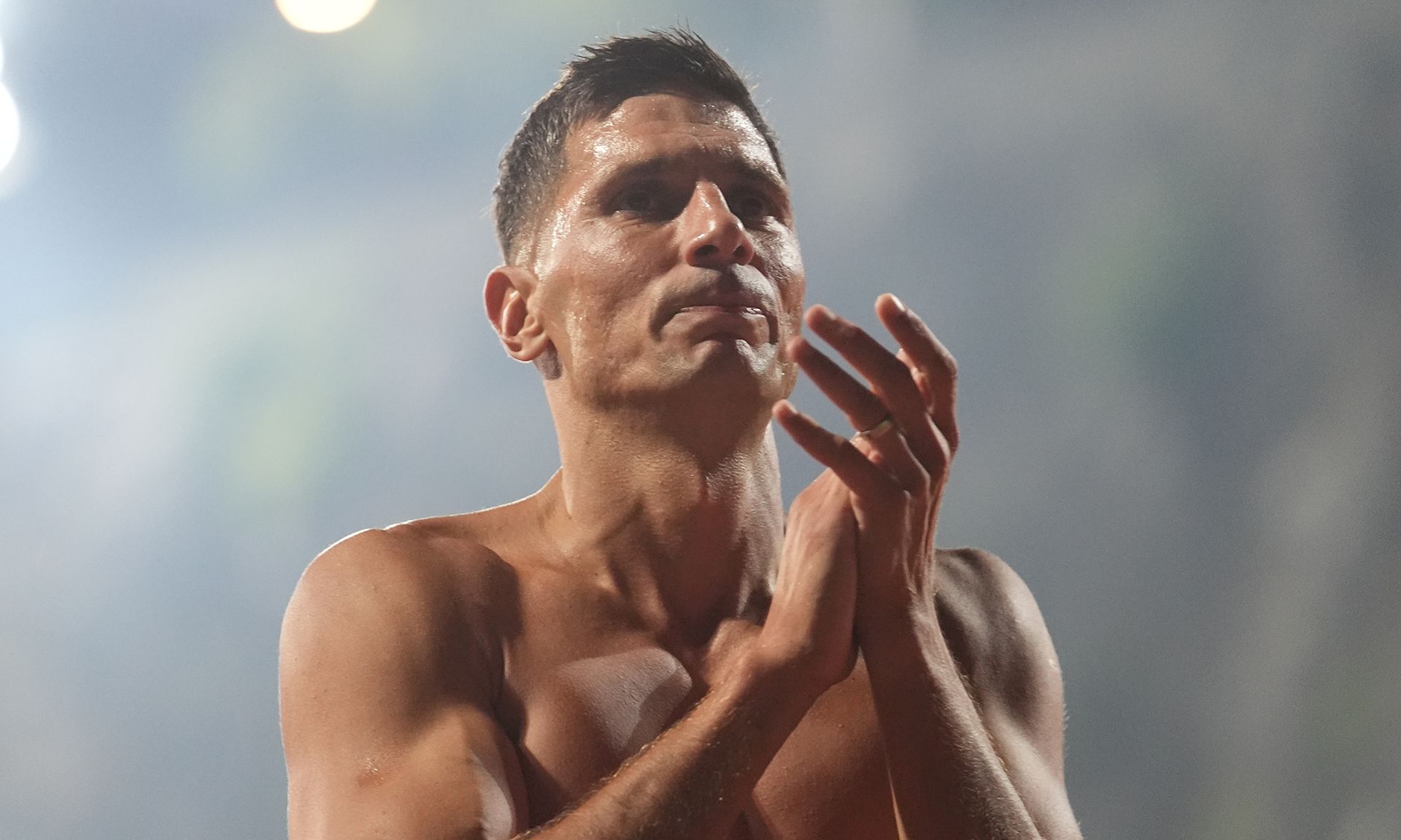 Ein athletischer Mann mit kurzen Haaren clappt, während er in einem sportlichen Umfeld steht. Er hat einen intensiven Ausdruck auf dem Gesicht.