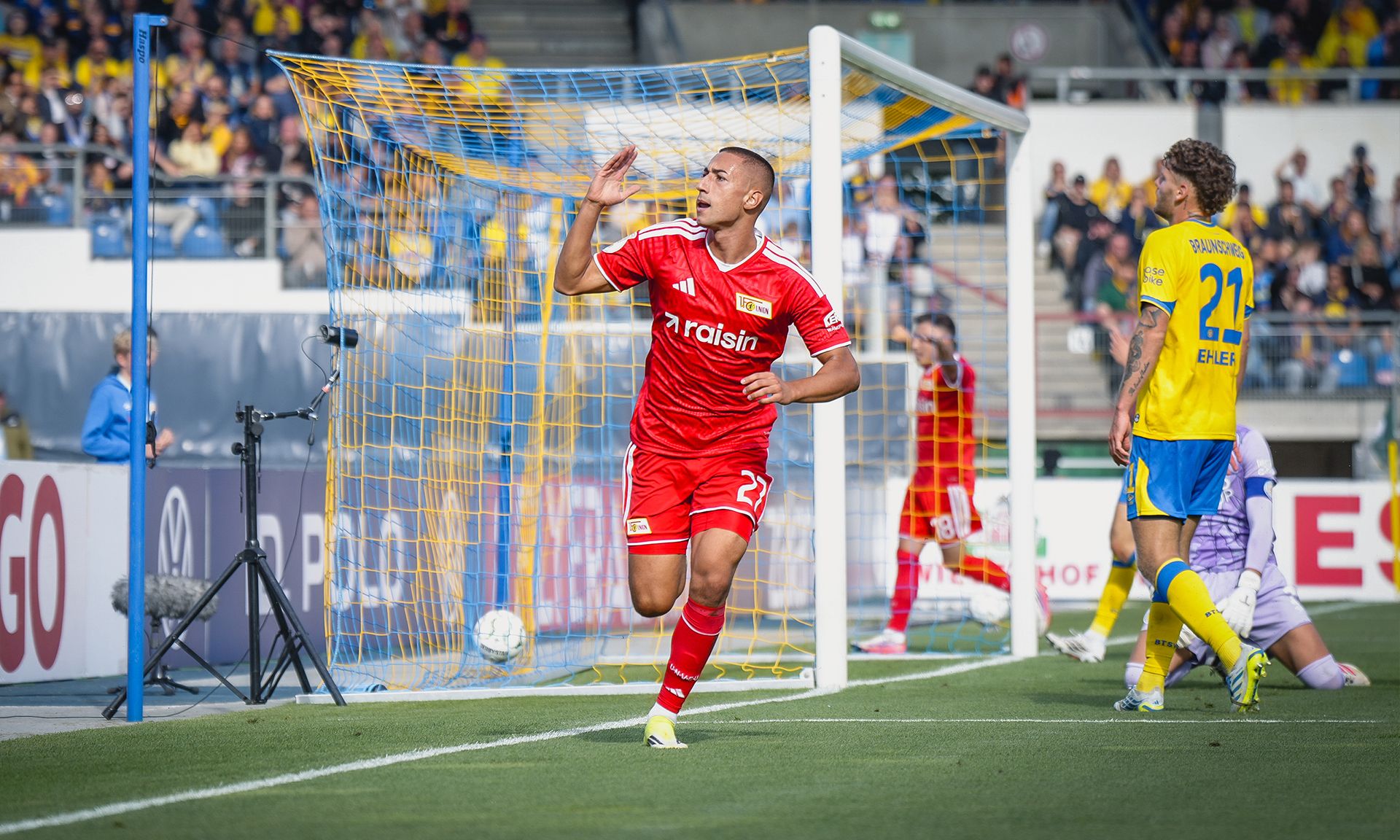 Ein Spieler in rotem Trikot jubelt nach einem Tor, während die gegnerische Mannschaft im Hintergrund steht.