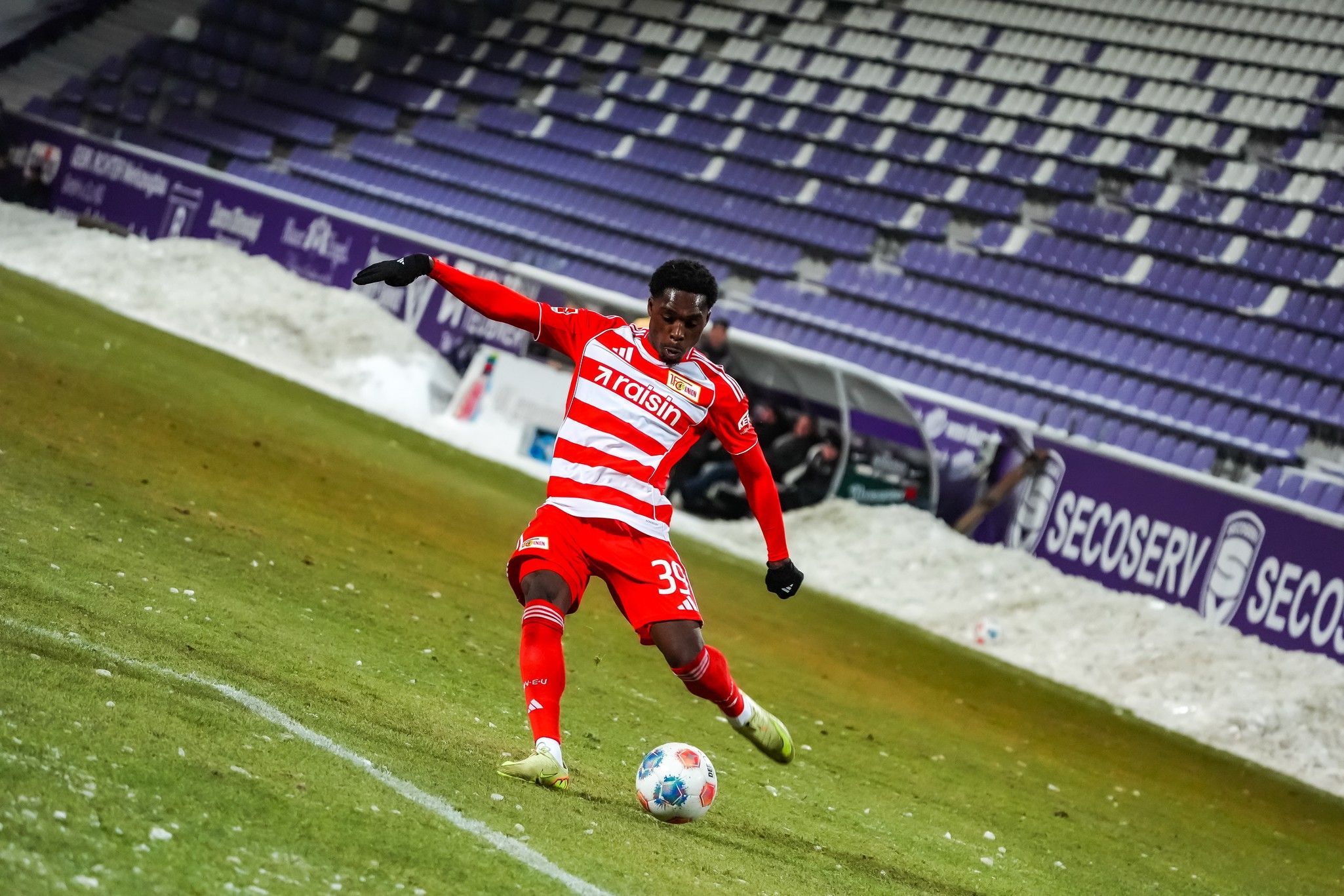 Spieler in rotem Streifen-Trikot tritt einen Fußball auf einem Rasenfeld mit schneebedeckten Stadionrängen im Hintergrund.