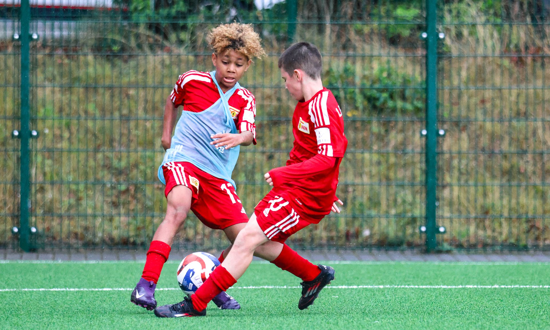 Zwei Kinder in roten Fußballtrikots spielen auf einem Kunstrasenfeld, während sie um den Ball kämpfen.