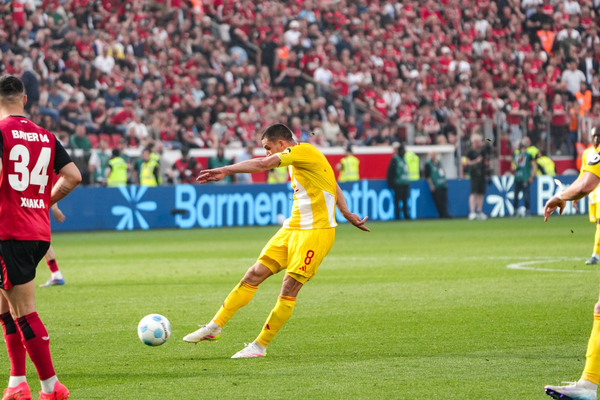 Spieler tritt den Ball während eines Fußballspiels, im Hintergrund jubelnde Fans in roten Trikots.