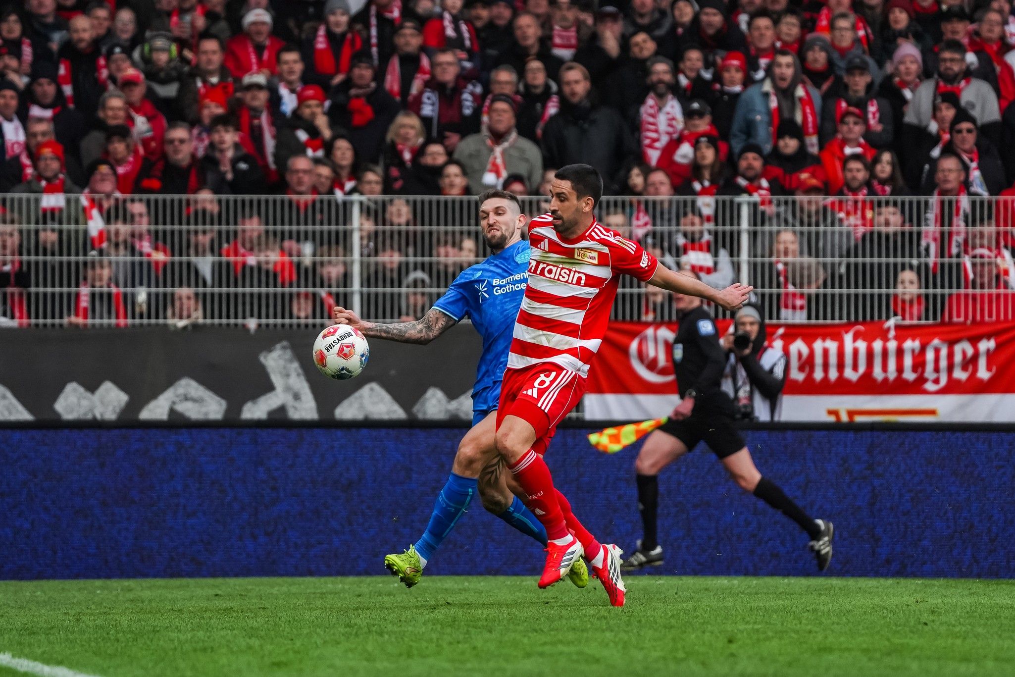 Spielszene: Ein Spieler in rot-weißem Trikot versucht, den Ball gegen einen blauen Spieler zu kontrollieren, während die Zuschauer jubeln.