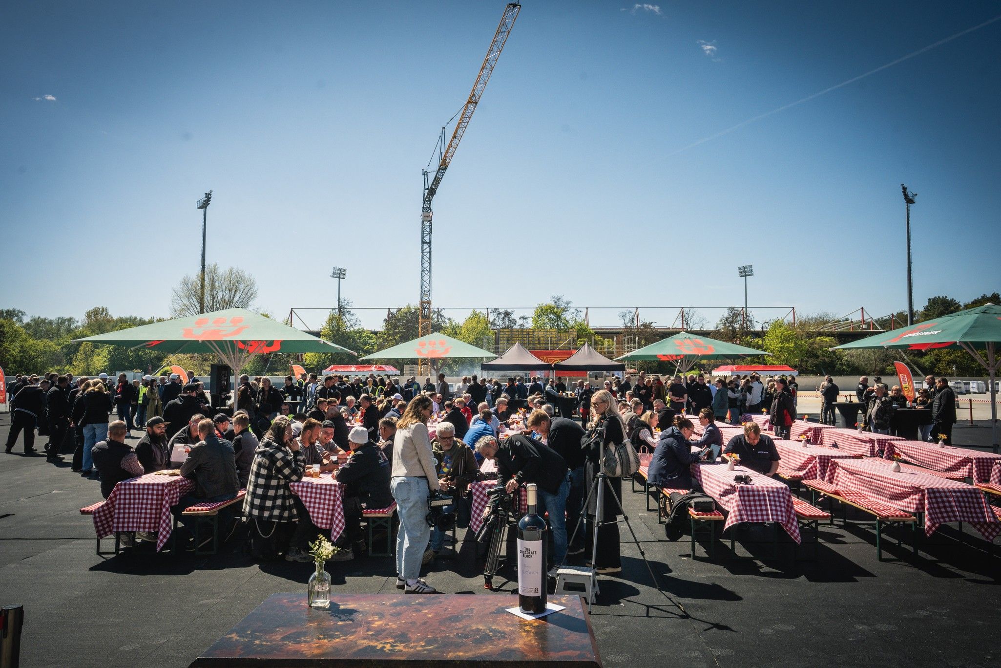 Menschenmenge bei einem Outdoor-Event mit Tischen unter Sonnenschirmen und einer klaren blauen Himmel.