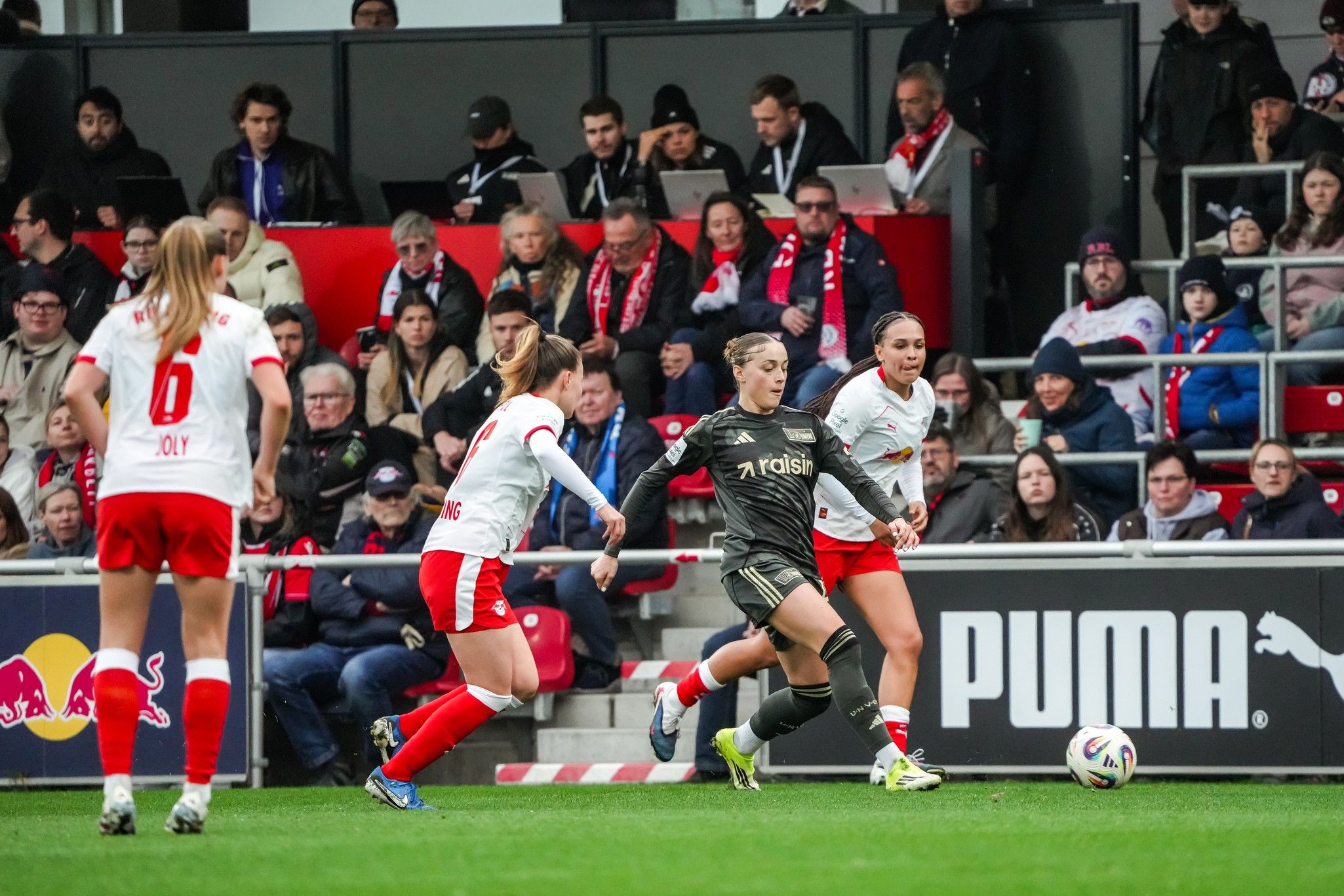 Spielszene im Frauenfußball: Eine Spielerin dribbelt den Ball, während zwei Gegnerinnen versuchen, ihn zu erobern. Zuschauer im Hintergrund.