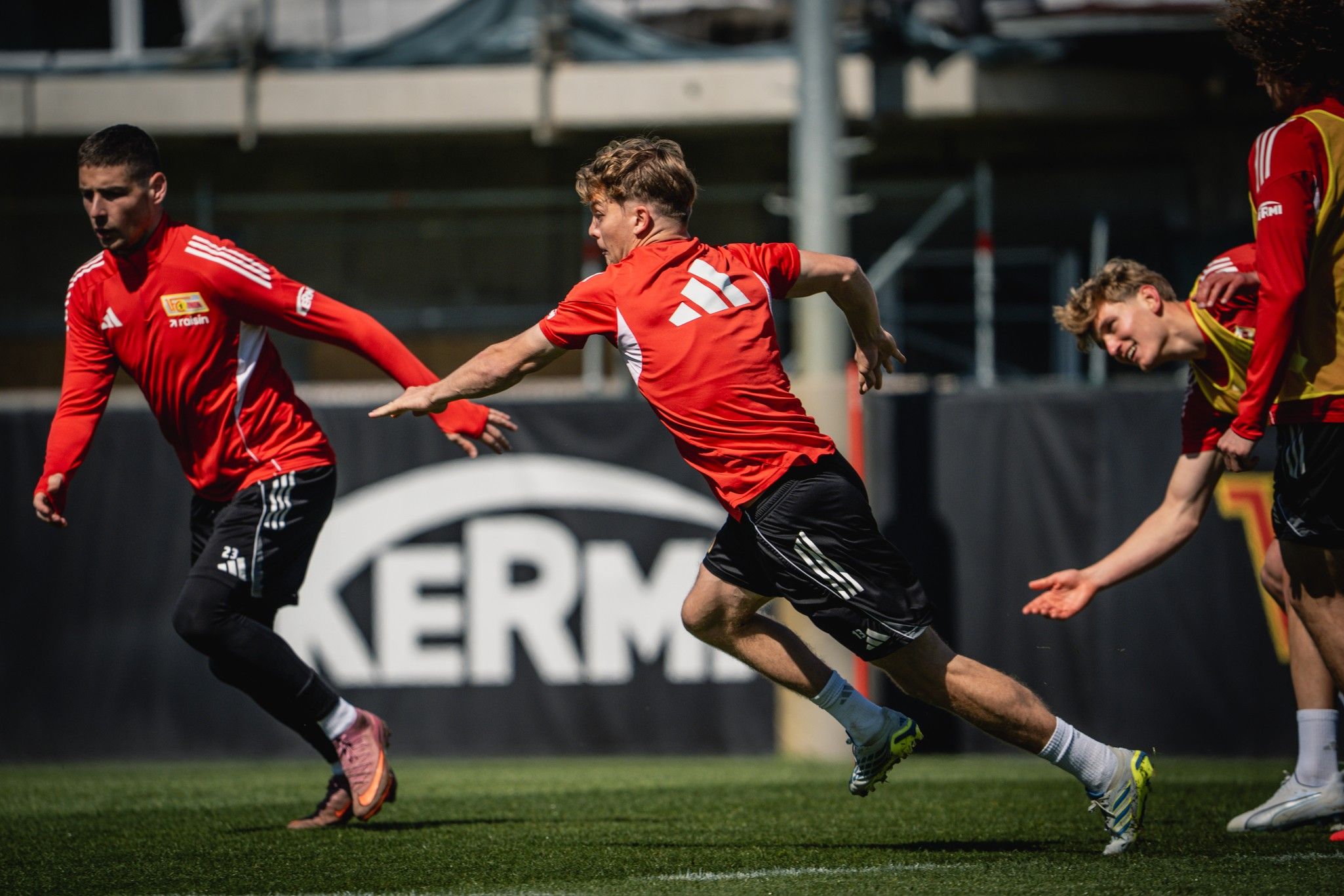 Spieler in roten Trikots trainieren auf einem Fußballfeld, während einer sprintet und die anderen in verschiedenen Aktionsstellungen sind.