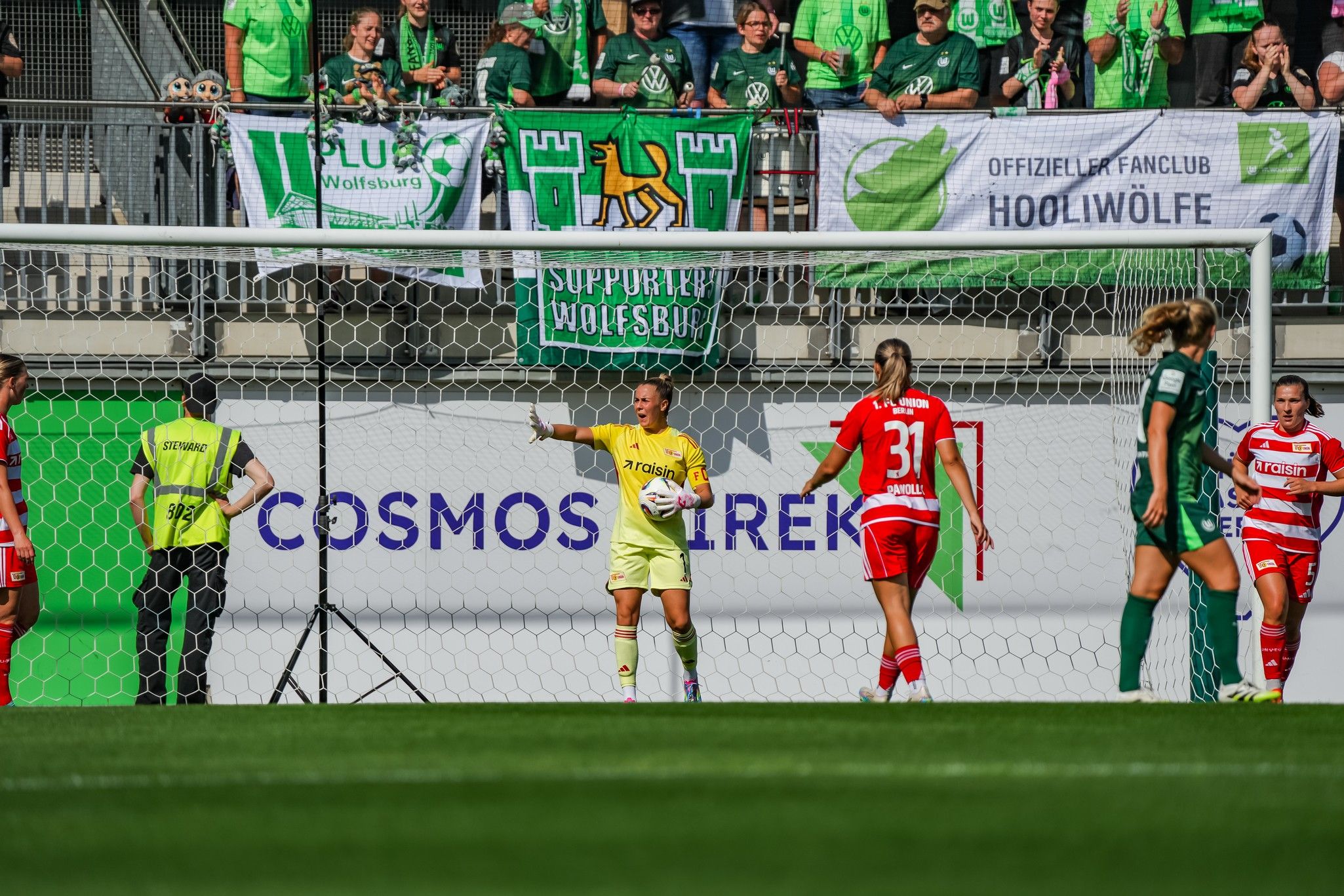 Torwart im oberen Bereich des Bildes signalisiert mit einer Hand, während Spielerinnen eines Spiels auf dem Feld sind. Fans im Hintergrund.