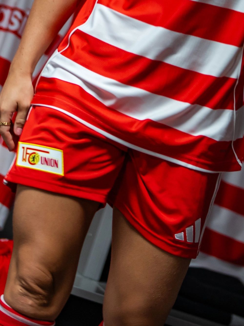 Rote Sportshorts mit weißen Streifen und einem Logo von "1. FC Union", getragen von einem Athleten. Hintergrund mit rotem und weißen Banner.