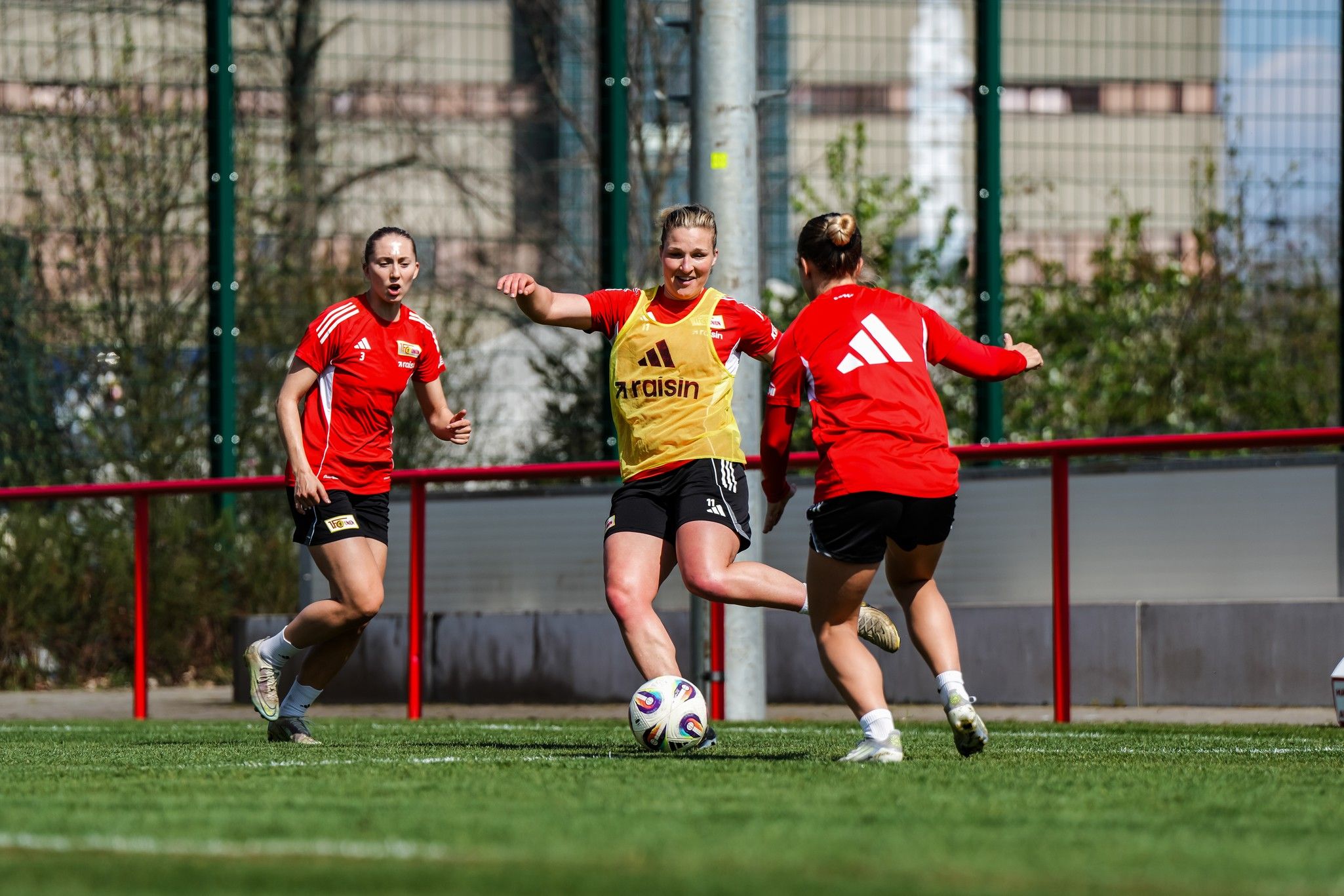 Drei Fußballspielerinnen trainieren auf einem Platz, eine Spielerin in gelber Weste schießt den Ball, während die anderen beiden reagieren.